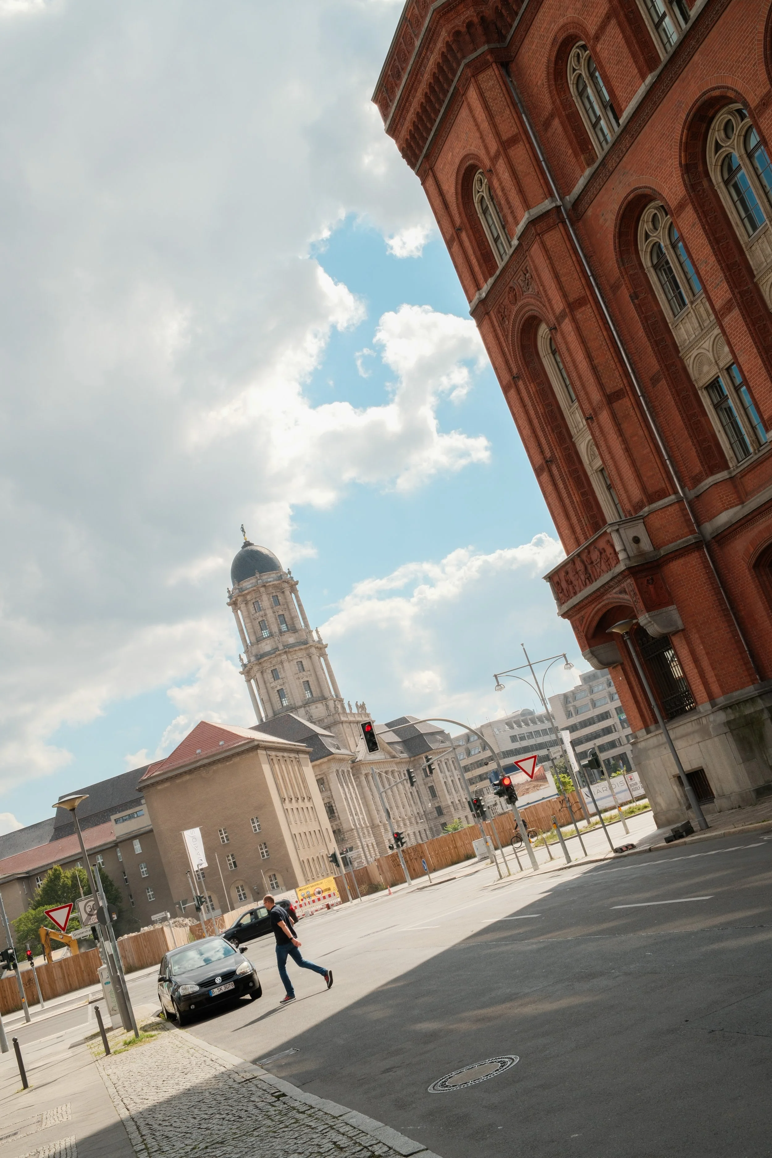 City street with red brick building, church with bell tower in background, traffic lights, a man walking across the street, and partly cloudy sky.