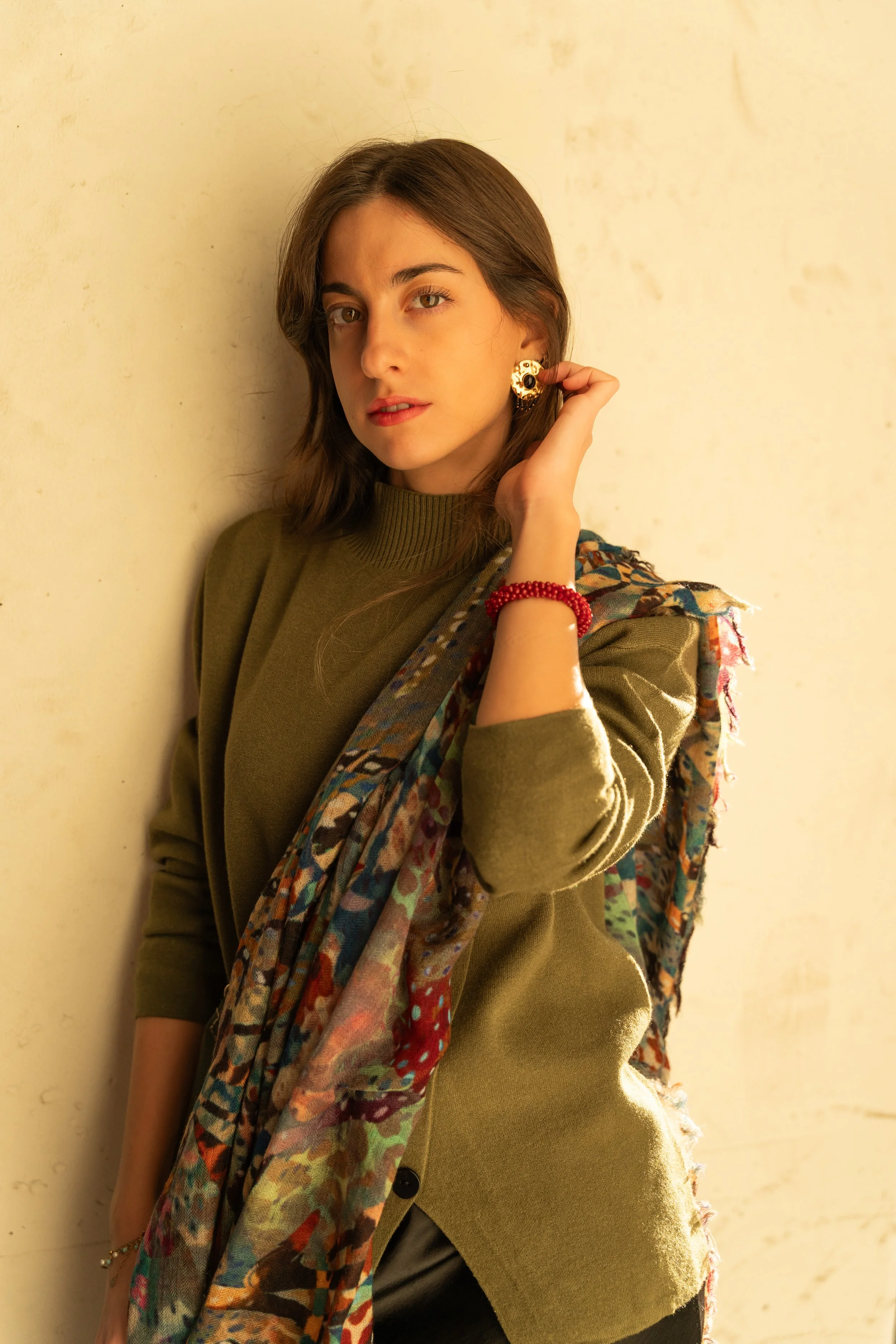 A young woman with long brown hair and light skin poses leaning against a beige wall, wearing a green sweater, colorful scarf, gold earrings, and a red bracelet.