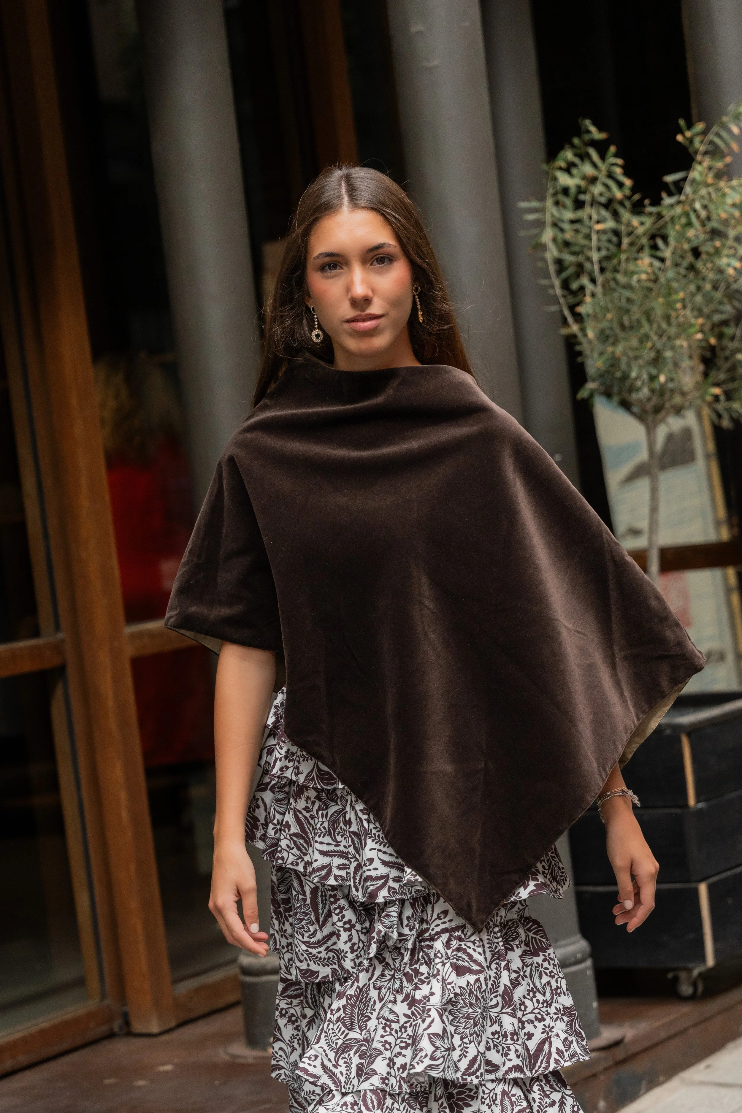 A young woman with brown hair, wearing earrings, a dark brown poncho, and a ruffled, floral black and white skirt, standing outside a building with glass doors and a potted plant.