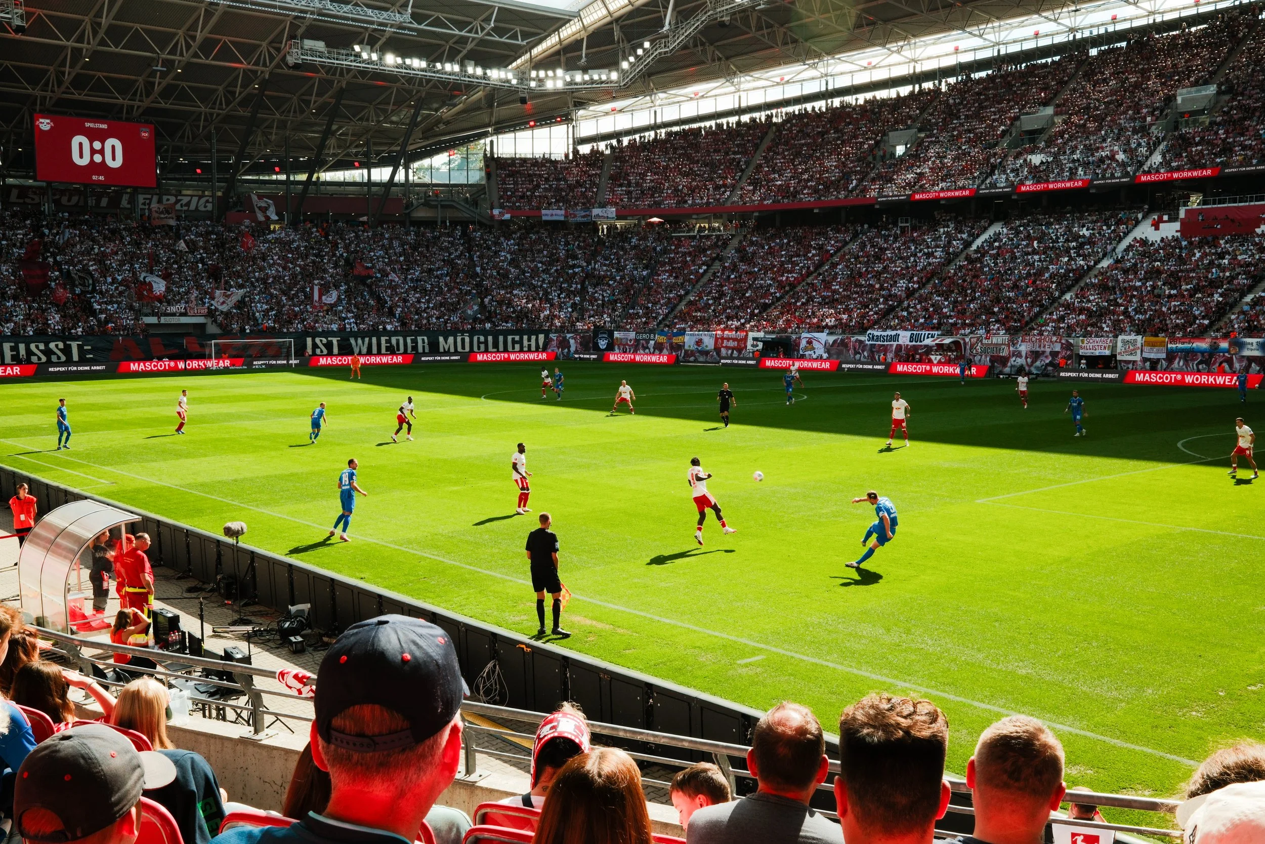 A soccer match in a large stadium filled with spectators, players on the field, and an electronic scoreboard showing 0:0.
