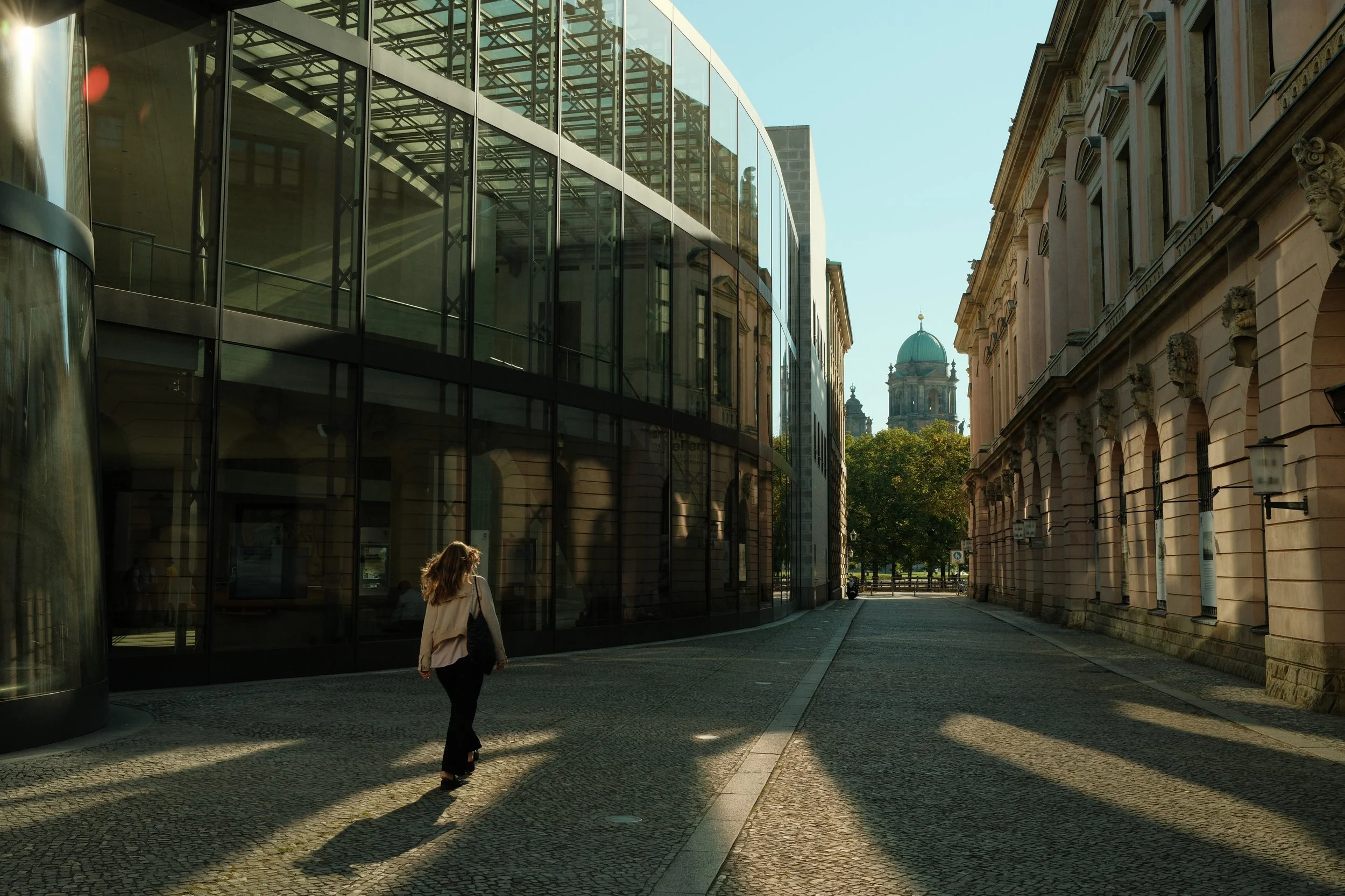 A woman walking along a cobblestone street in a city, with modern glass buildings on the left and historic stone buildings on the right, with a domed building in the background, and shadows cast by trees.