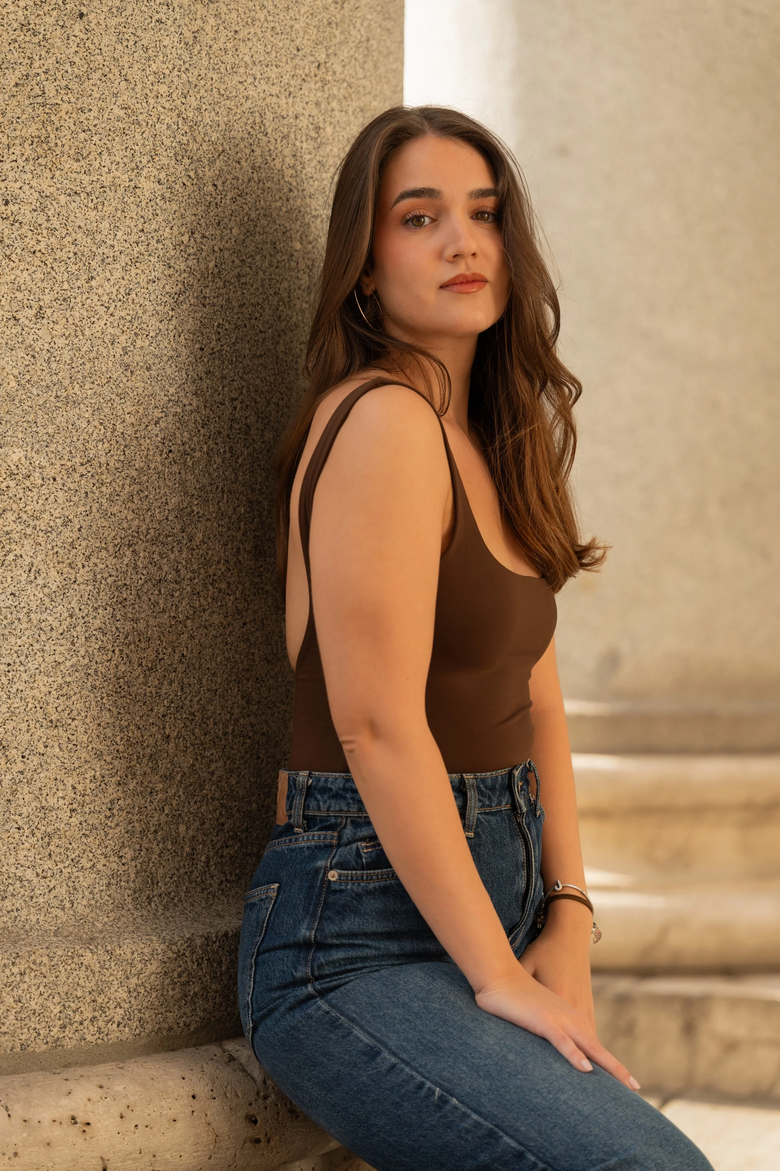A young woman with long brown hair, wearing a brown tank top and blue jeans, sitting on a low stone ledge against a textured concrete wall.