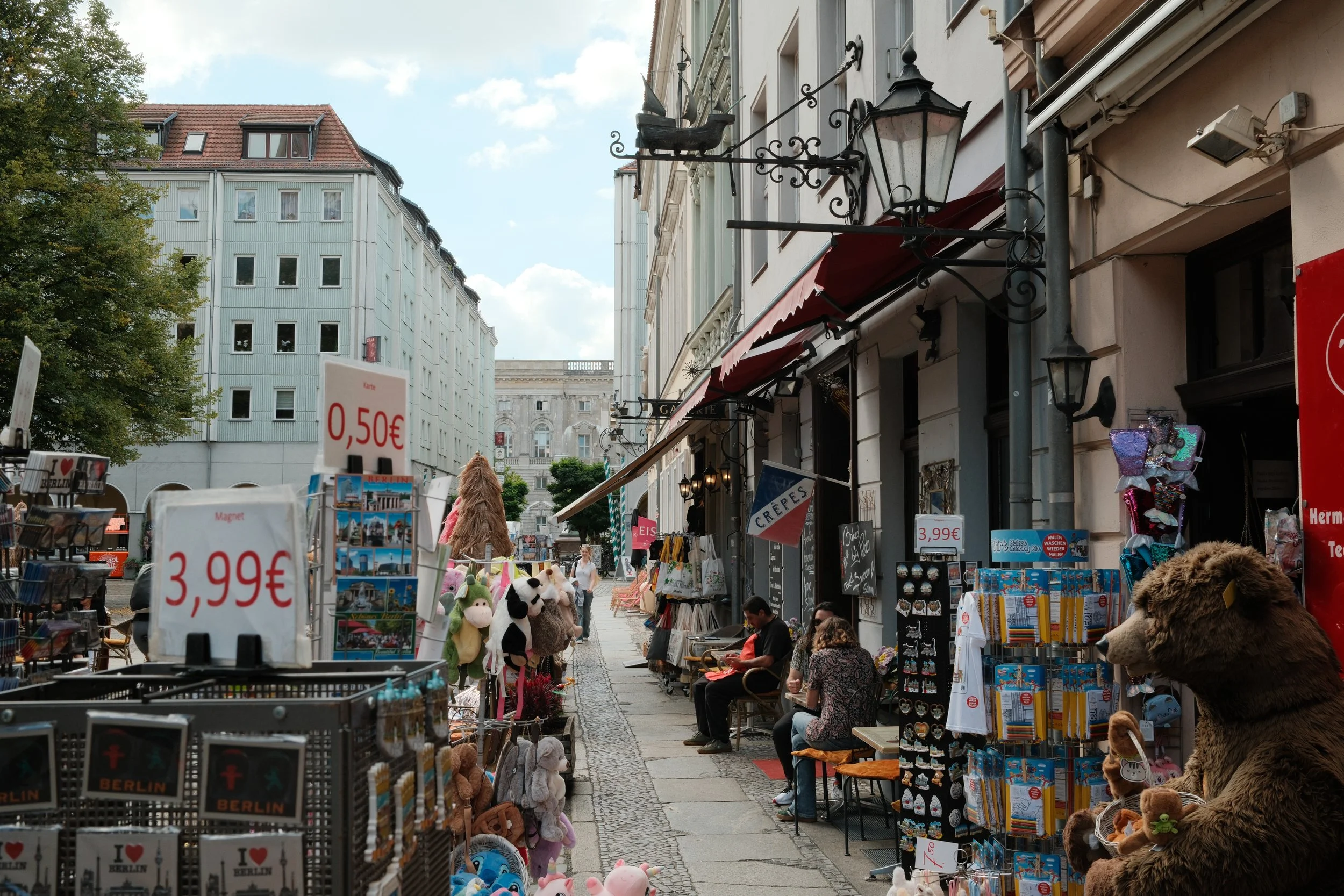 A street scene with souvenir stalls, stuffed animals, and people sitting outside cafés, with European-style buildings and a partly cloudy sky in the background.