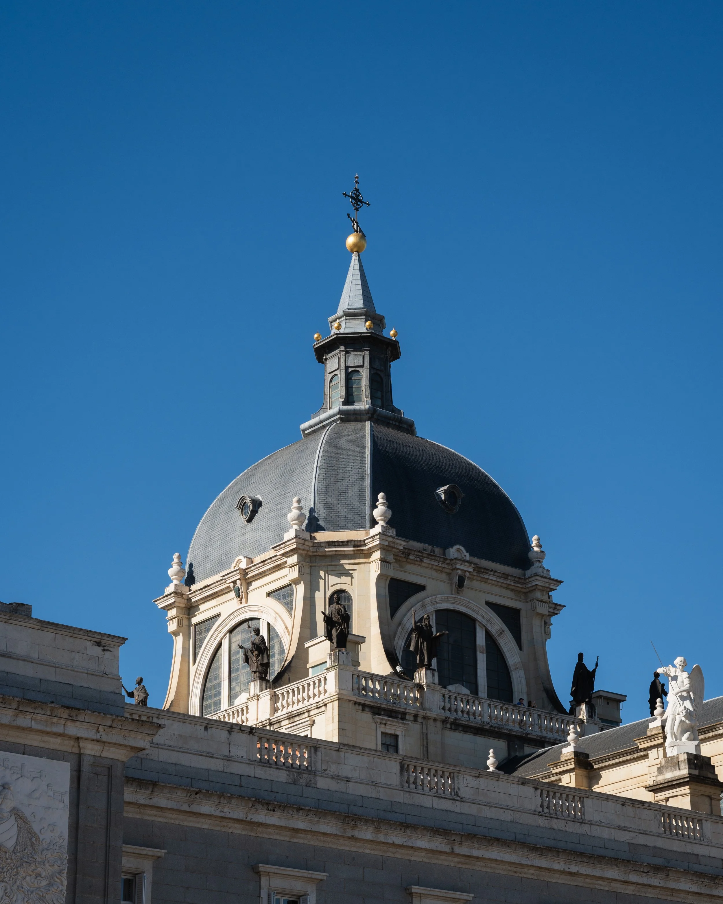 The upper part of a historic building featuring a decorative dome with statues and a weather vane atop a spire against a clear blue sky.