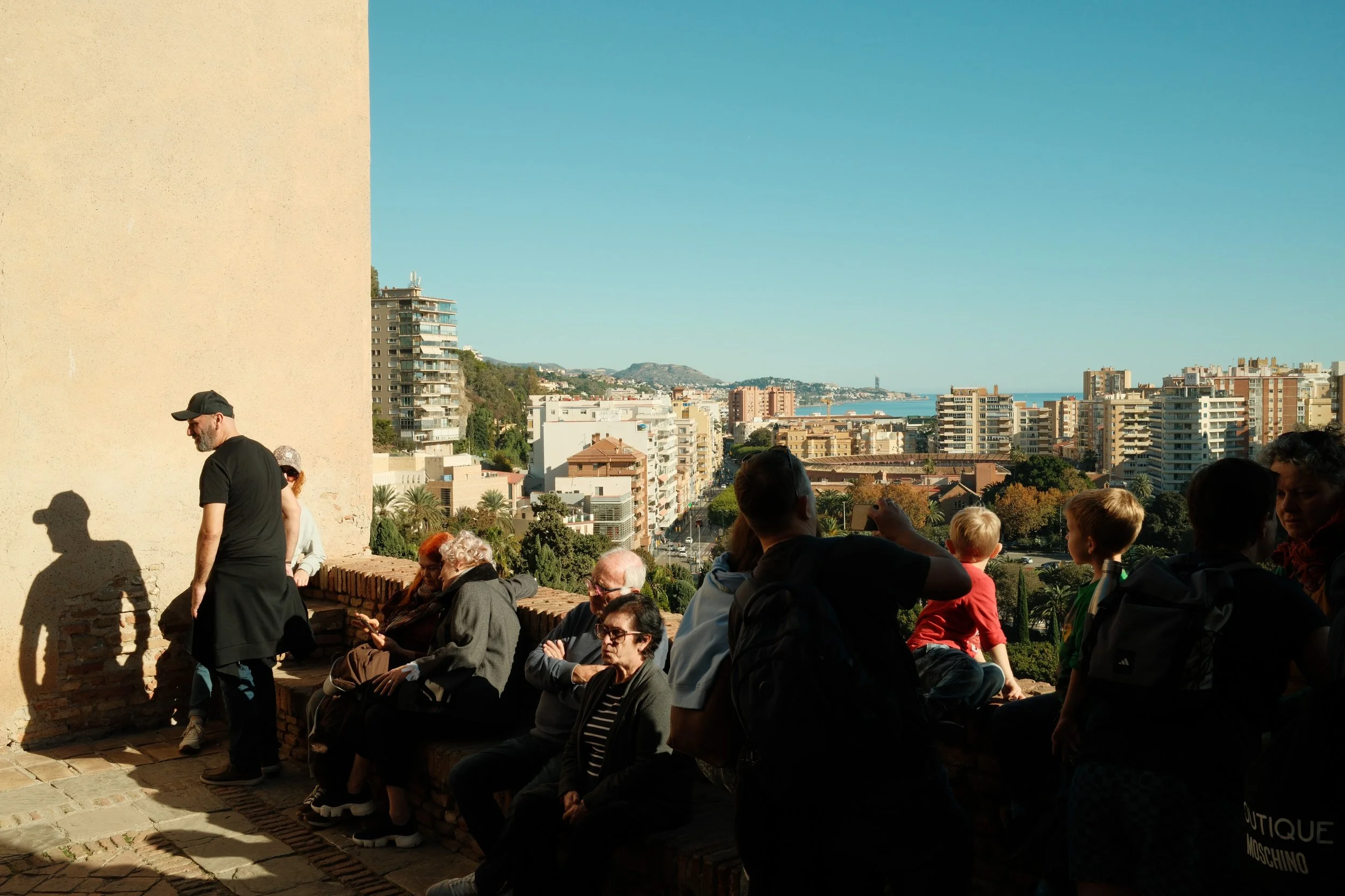 Group of people sitting and standing on a terrace overlooking a city with high-rise buildings and a distant coastline under a clear blue sky.