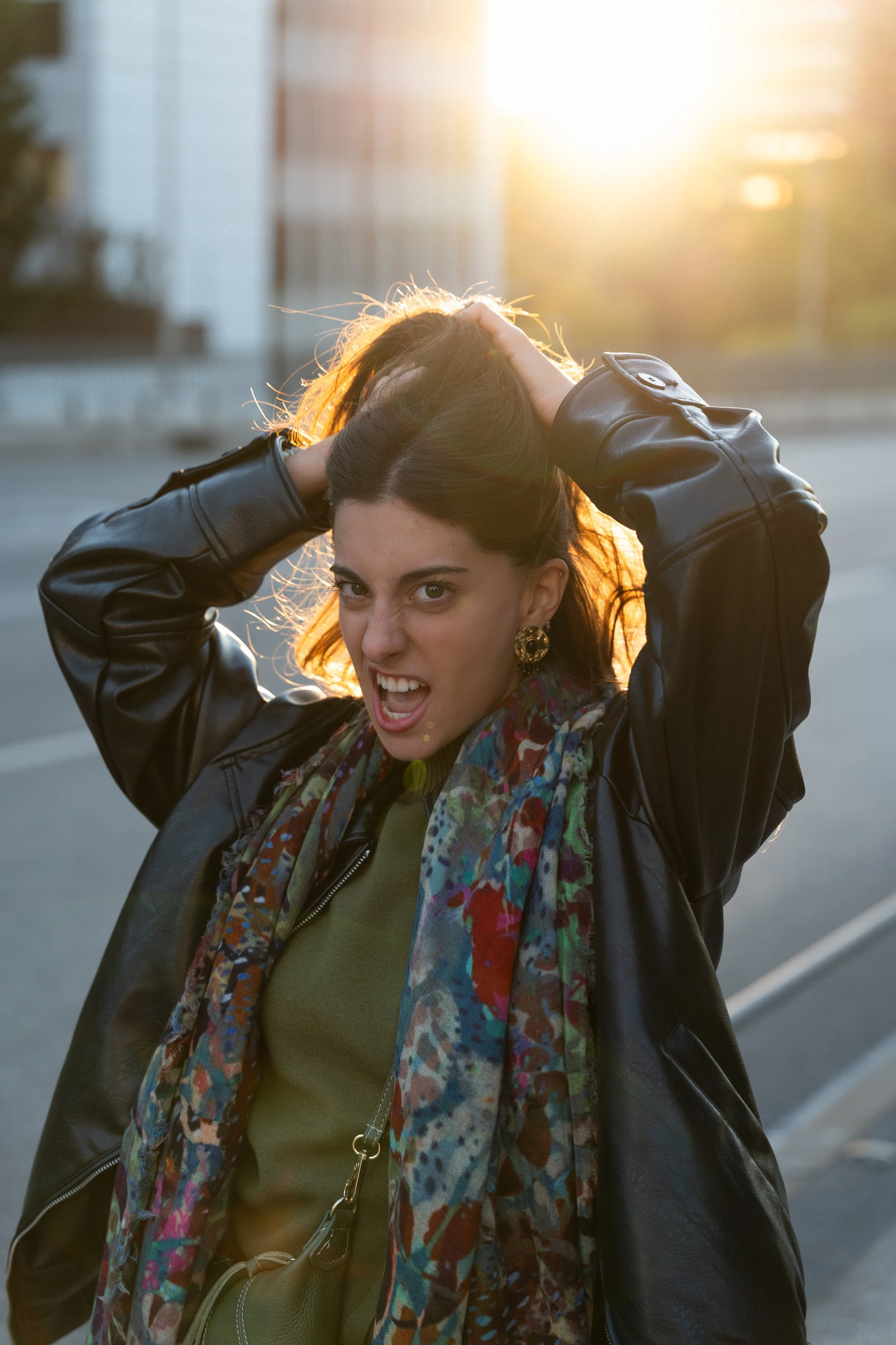 A woman with dark hair, wearing a leather jacket, colorful scarf, and gold earrings, making a playful, angry face with one eye squinted and hands on her head, standing outdoors at sunset.