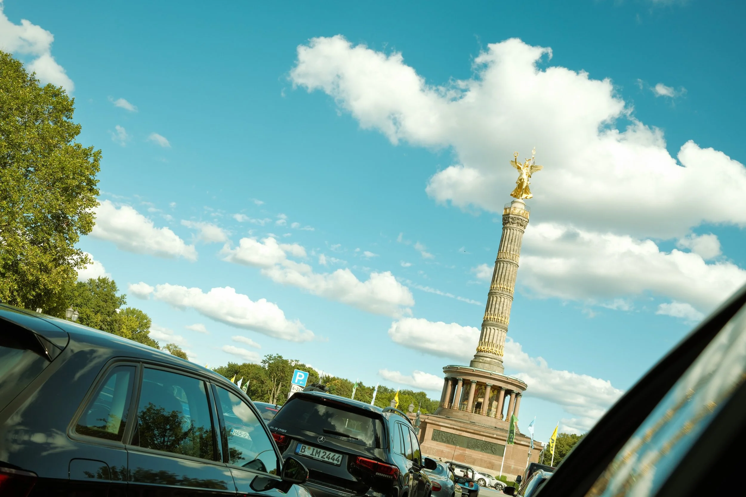A bronze statue of a woman with wings standing on top of a tall, decorated column with a circular base, surrounded by trees and parked cars, under a partly cloudy blue sky.