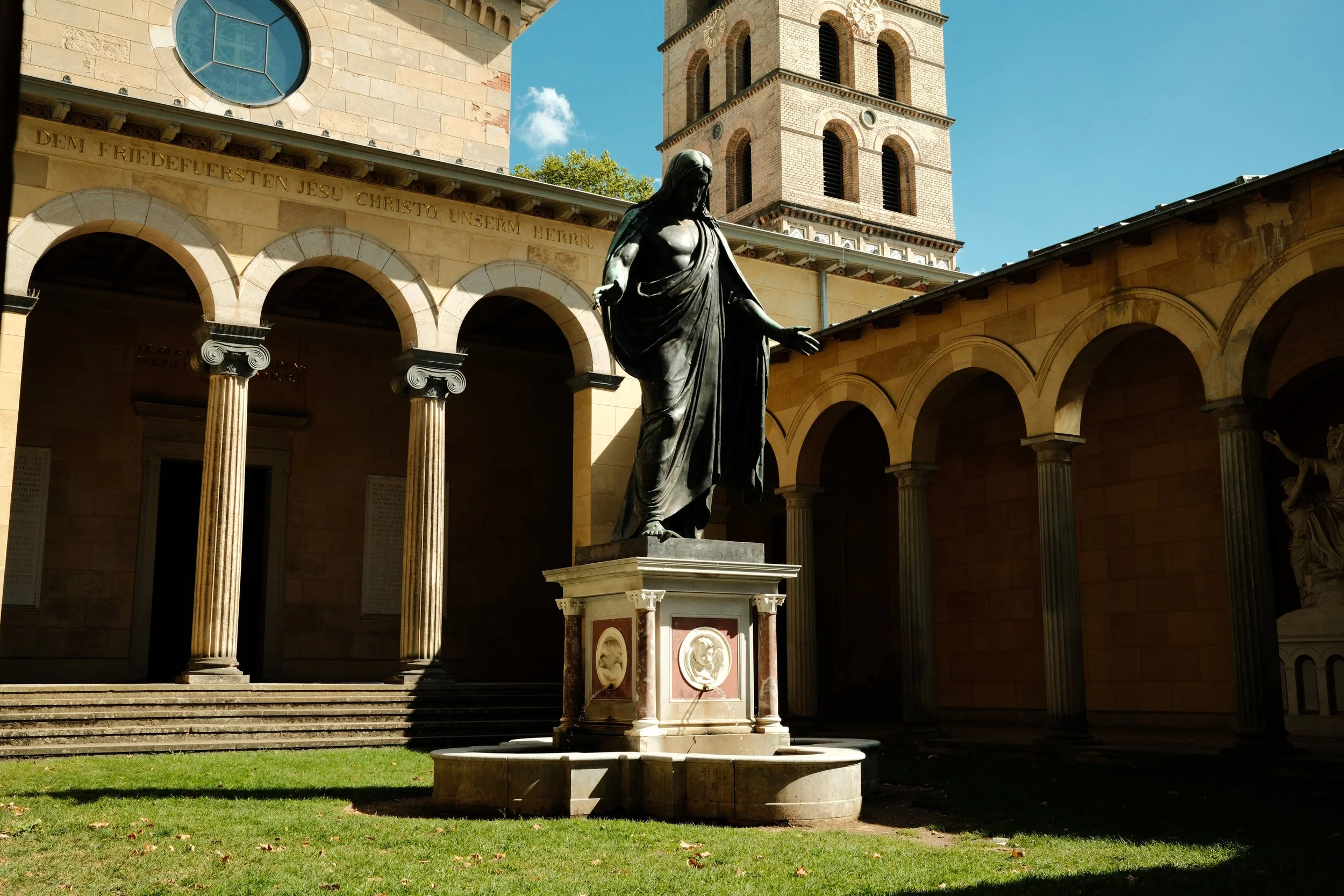 A bronze statue of Jesus Christ standing in a courtyard with arched colonnades, steps, and a tower in the background, under a blue sky with clouds.