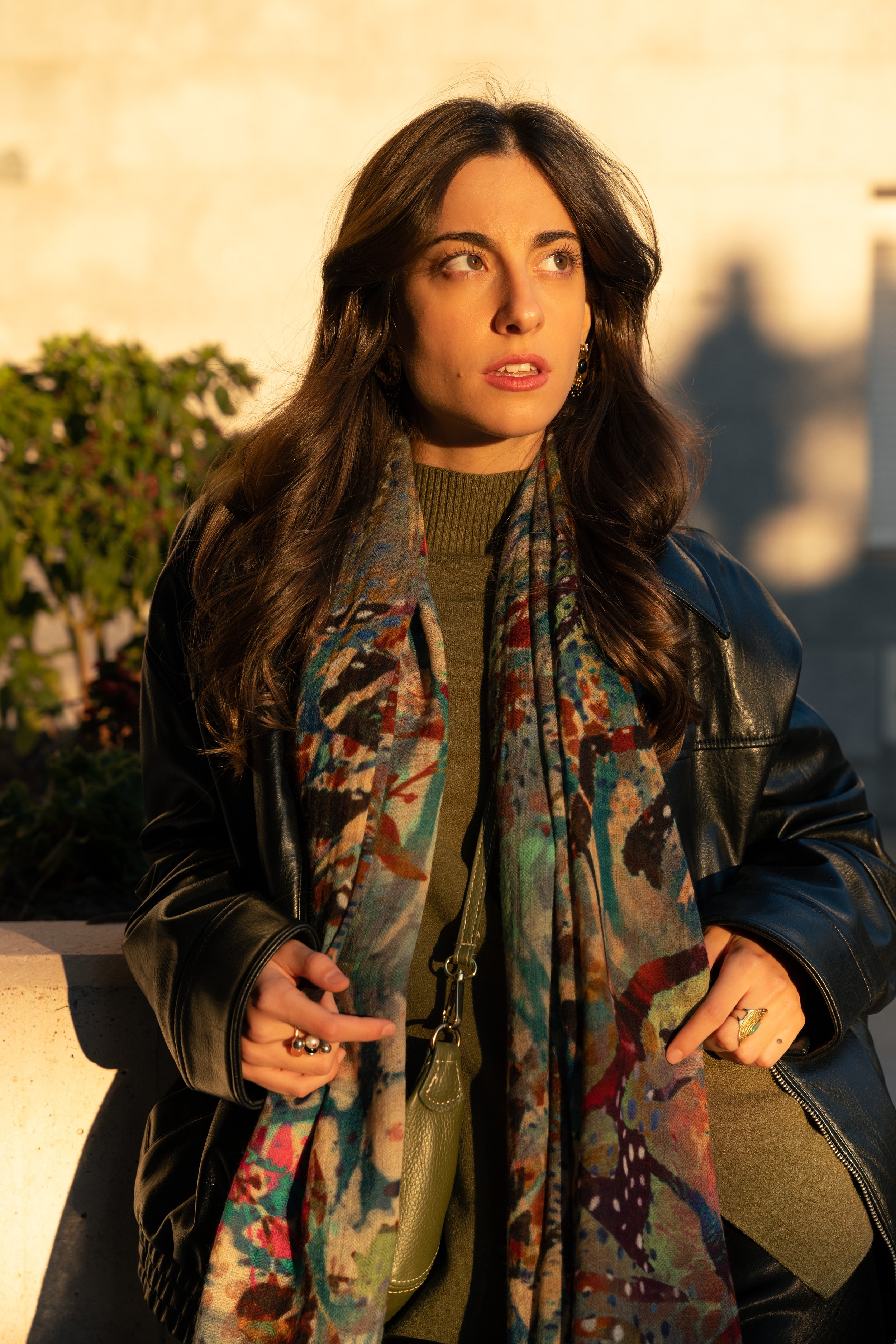 A woman with long wavy brown hair, wearing a black leather jacket, green shirt, a colorful patterned scarf, and gold jewelry. She is outdoors, illuminated by warm sunlight, standing near a low wall with bushes in the background.