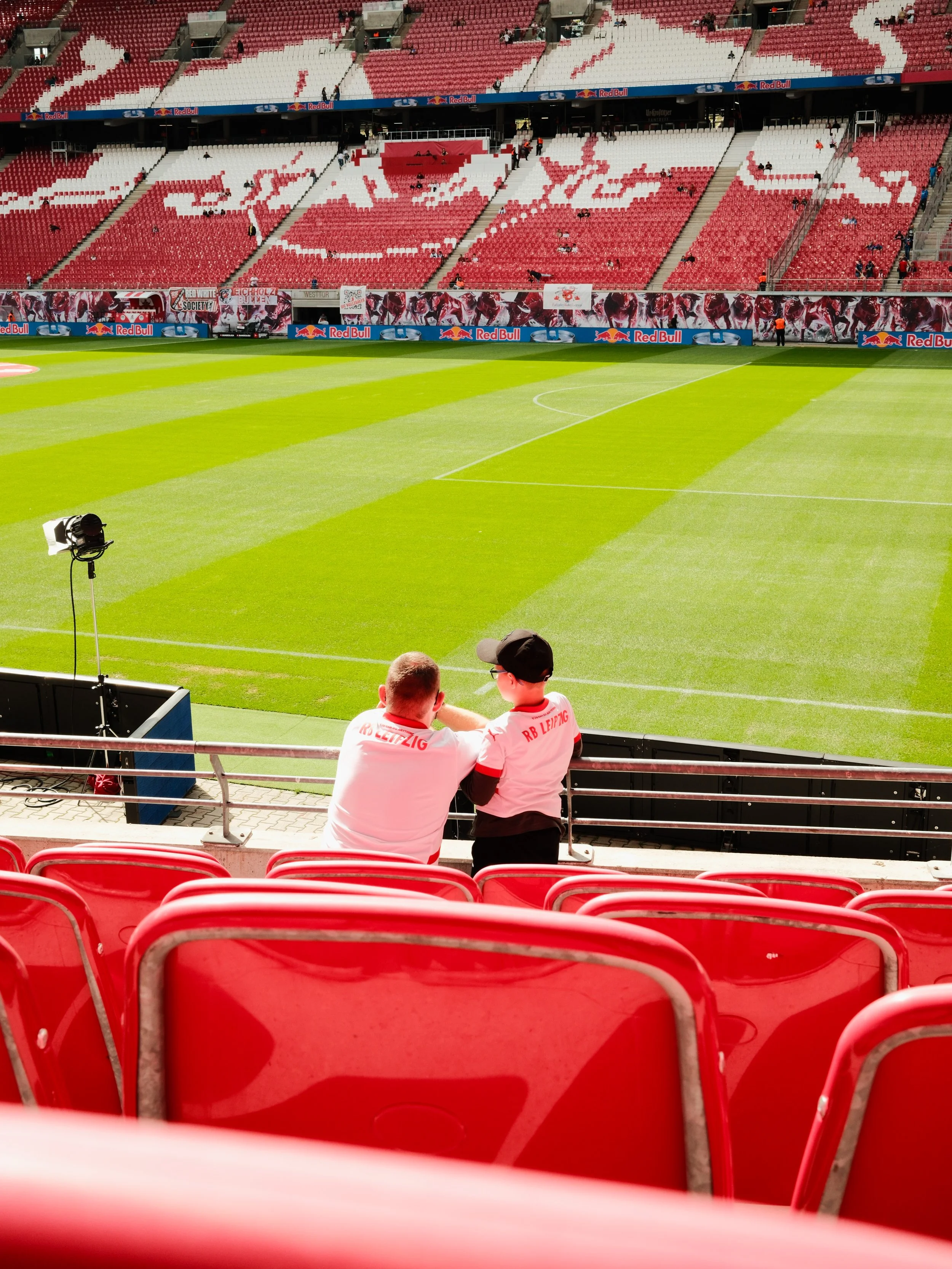 Two fans sitting in the stands at a soccer stadium, watching the empty soccer field with green grass. The stadium seats are red, and there is advertising along the perimeter of the field, including Red Bull logos. The two fans are wearing white shirt