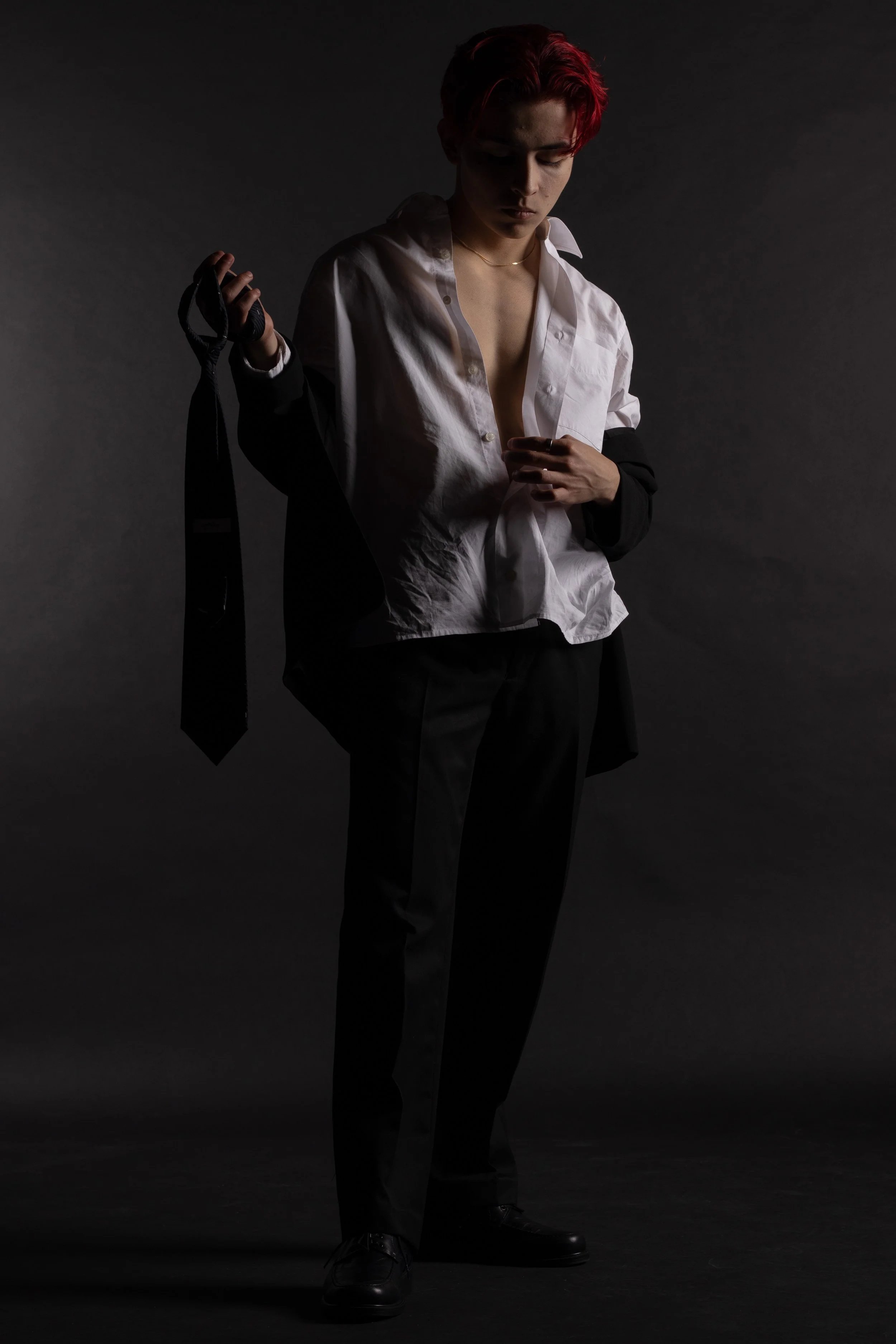 Person with red hair in a partially open white shirt, holding a black tie, in a dark studio setting.