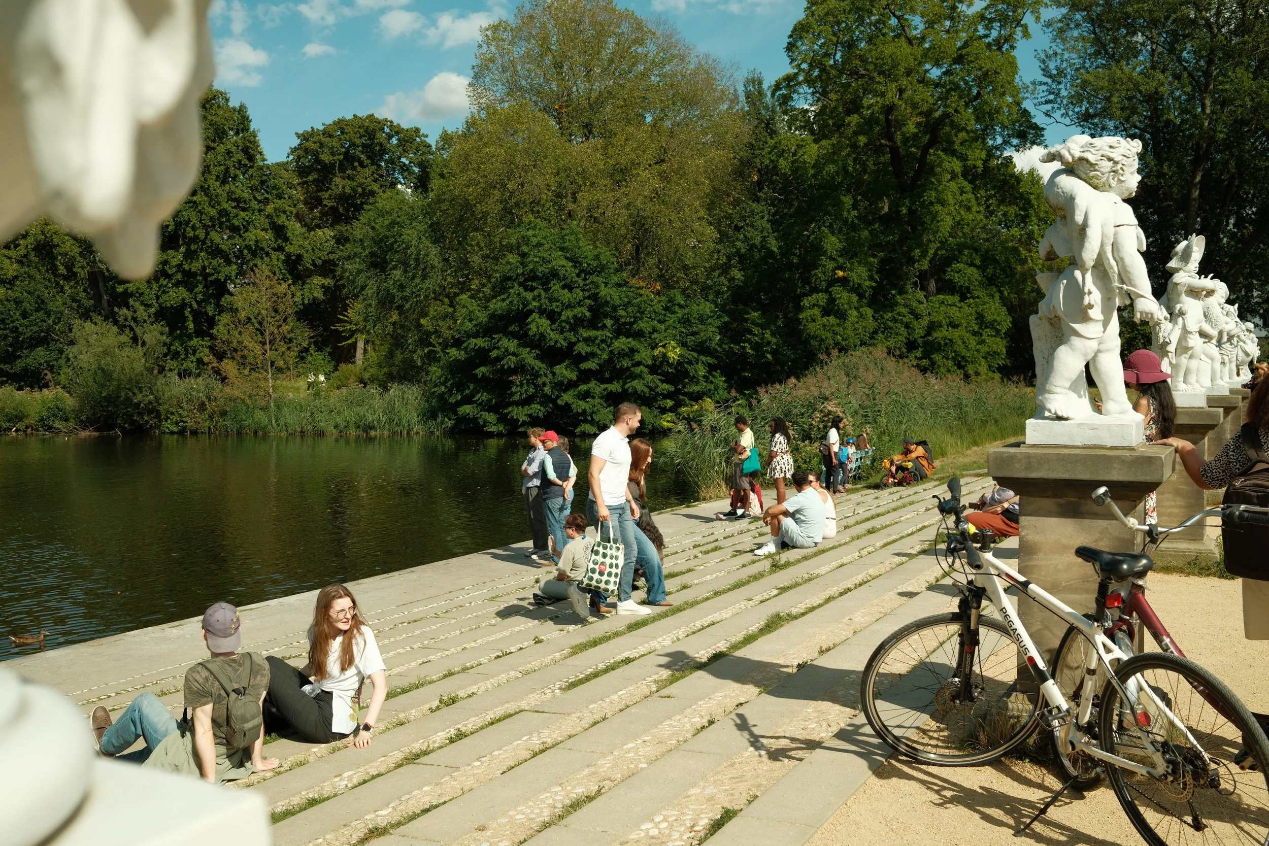 People sitting and walking along a lakeside path with statues and lush green trees in the background on a sunny day.