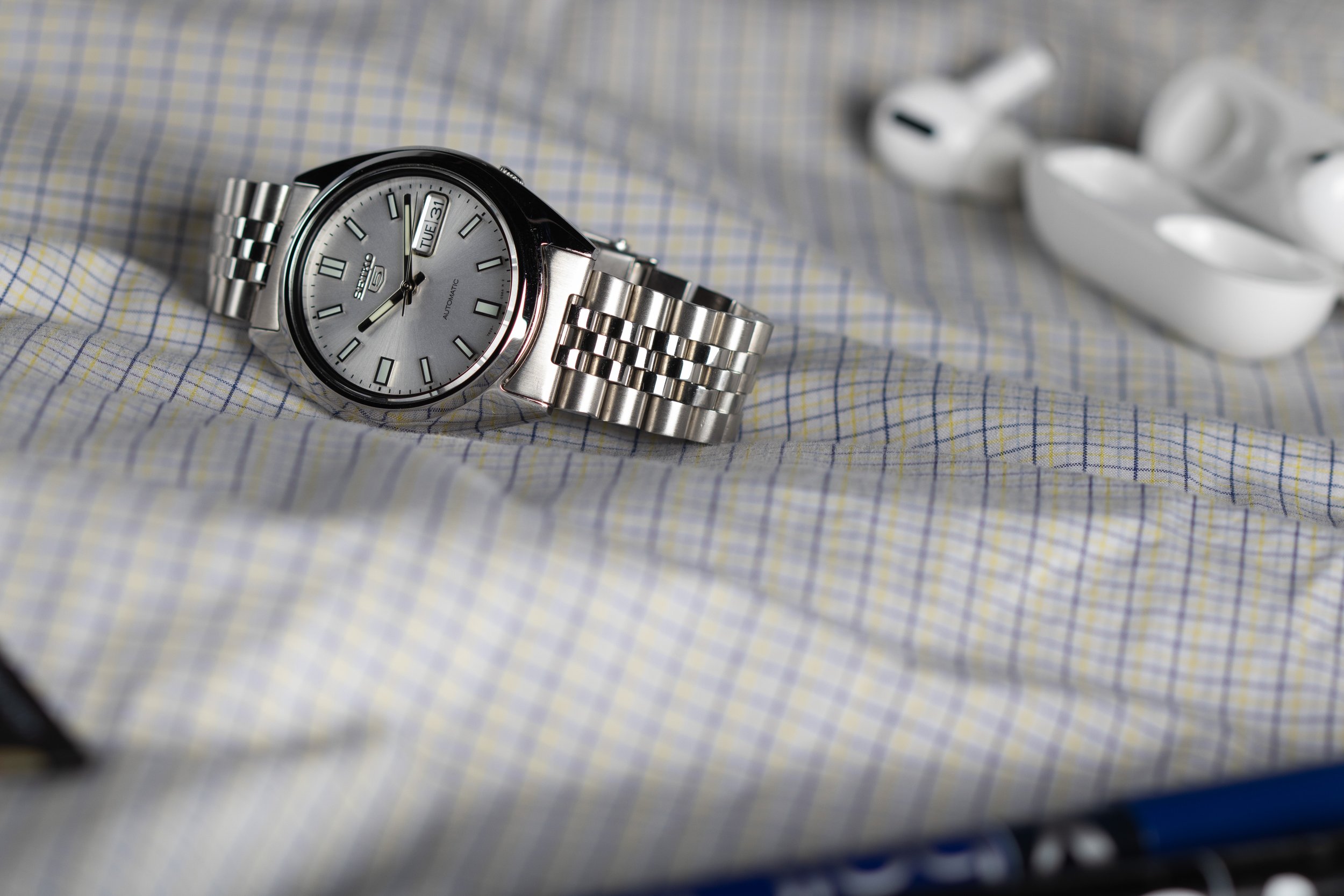 A silver Seiko wristwatch with a metal band on a checkered dress shirt, with a white wireless earbuds case in the background.