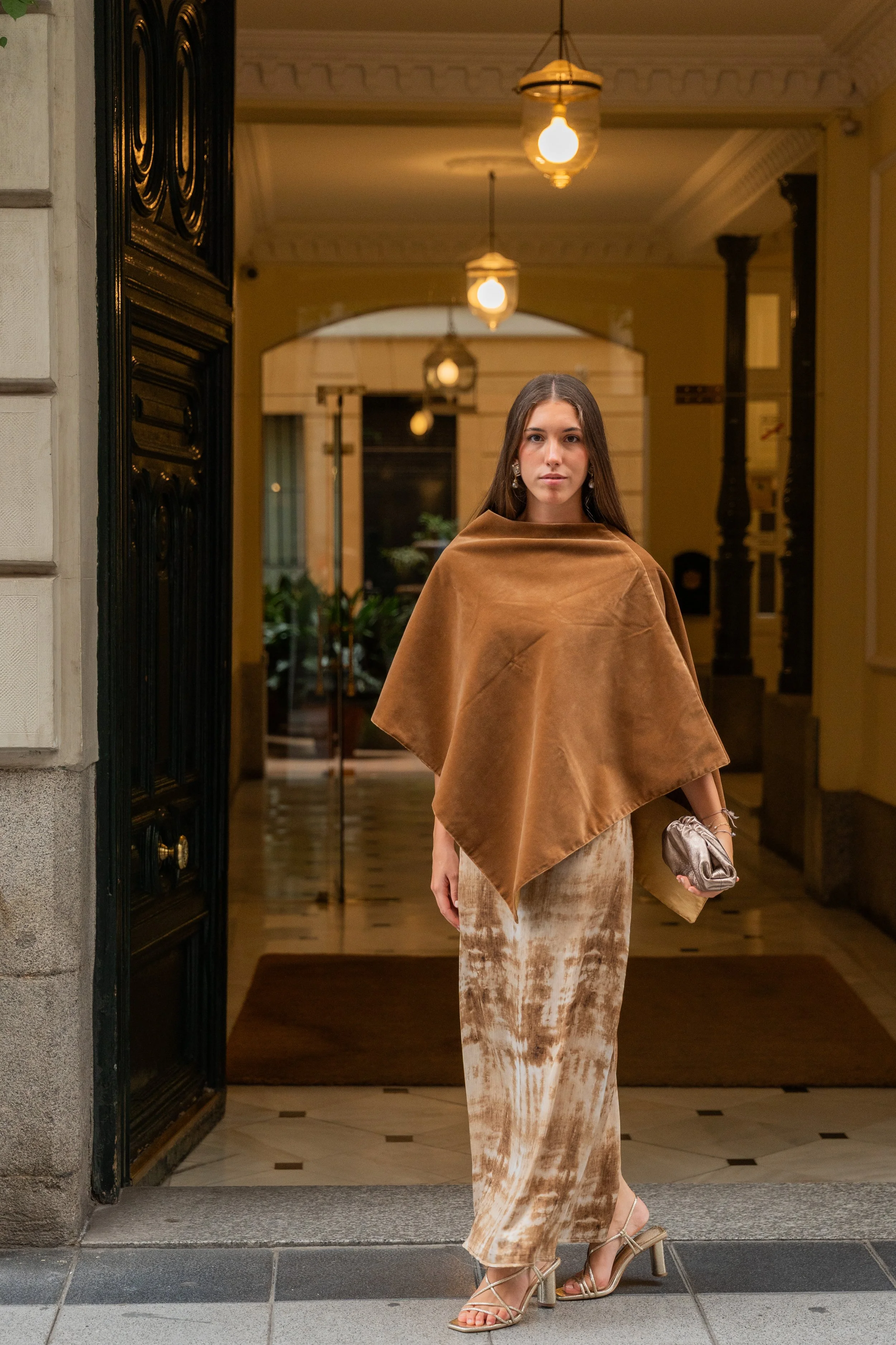A woman standing in an open doorway of a building, wearing a brown velvet shawl, a beige tie-dye skirt, and high-heeled sandals, holding a small silver clutch bag.