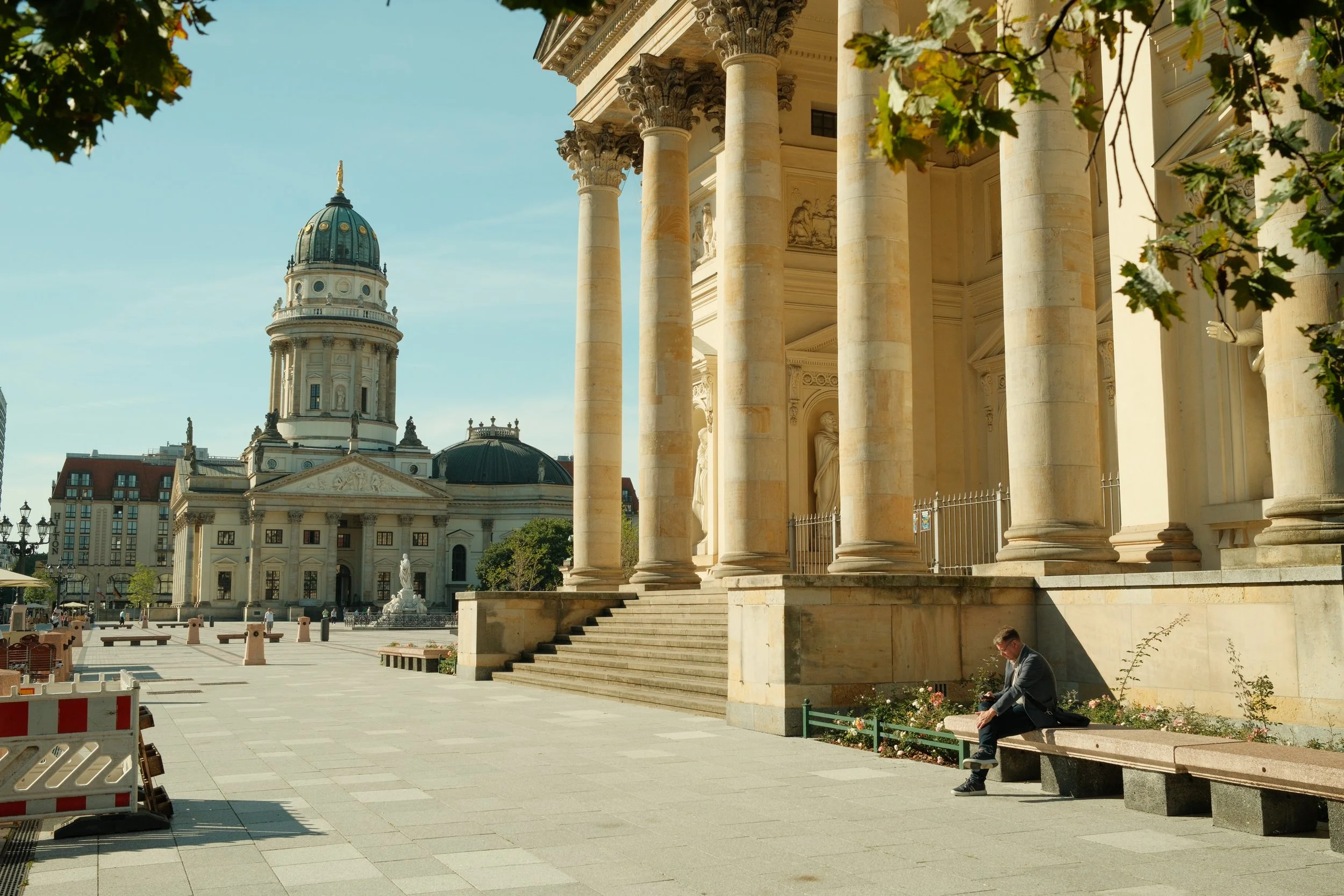 A man sitting on a bench in front of a large, ornate building with columns. In the background, there is a domed building with a statue on top, and the area appears to be a plaza with a few other structures and trees.