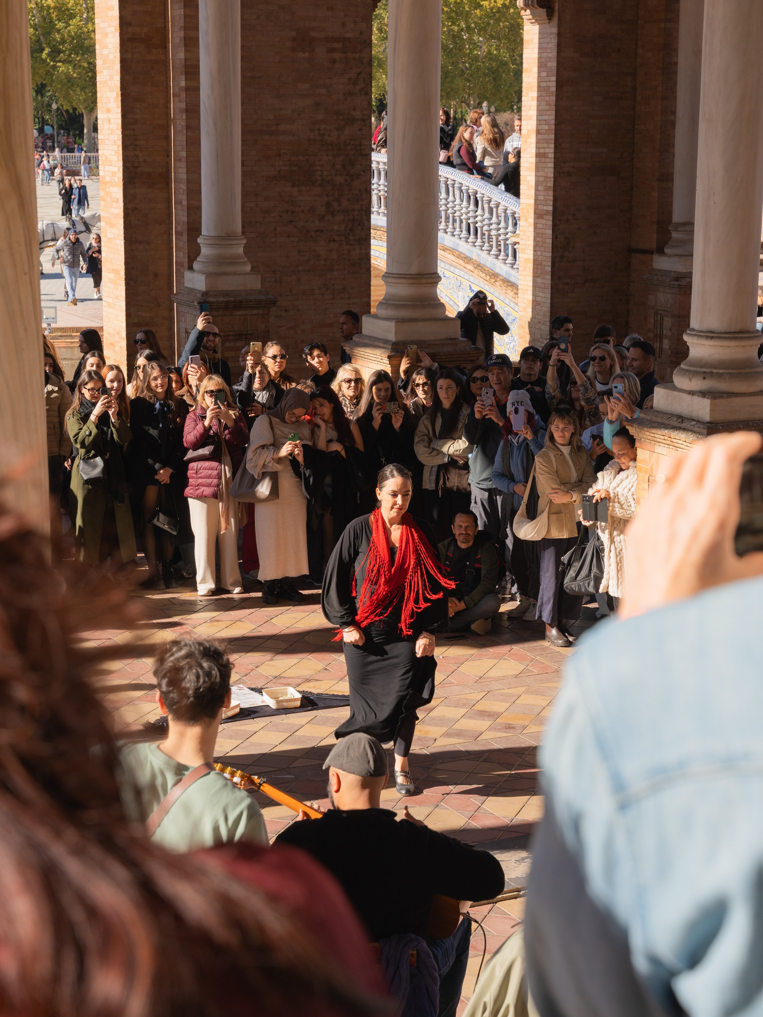 A performer with dark hair and a red scarf is dancing in front of a crowd gathered in a historic outdoor location with brick pillars and columns.