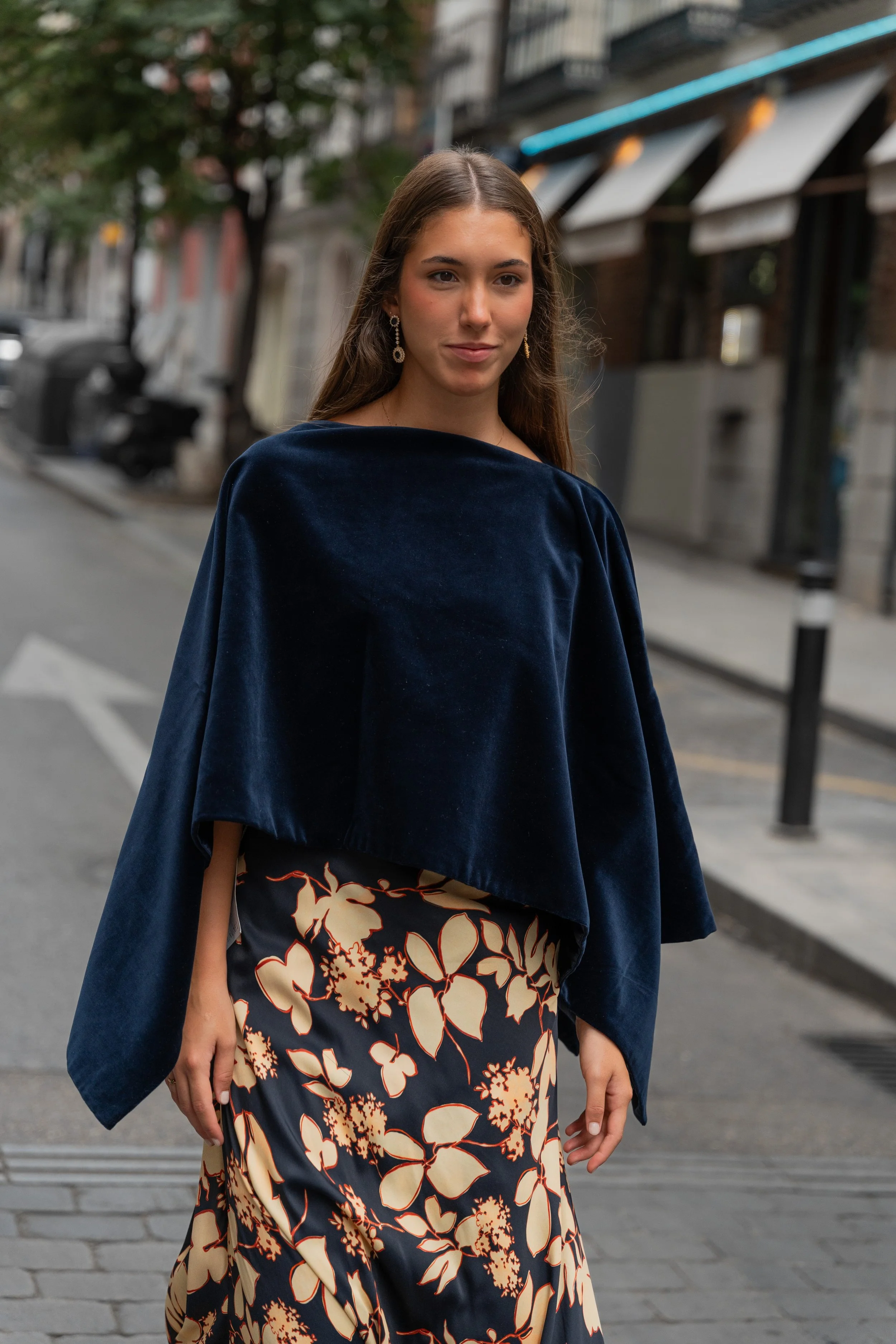 A young woman walking on a city street, wearing a navy blue velvet top and a floral printed skirt, with trees and buildings in the background.
