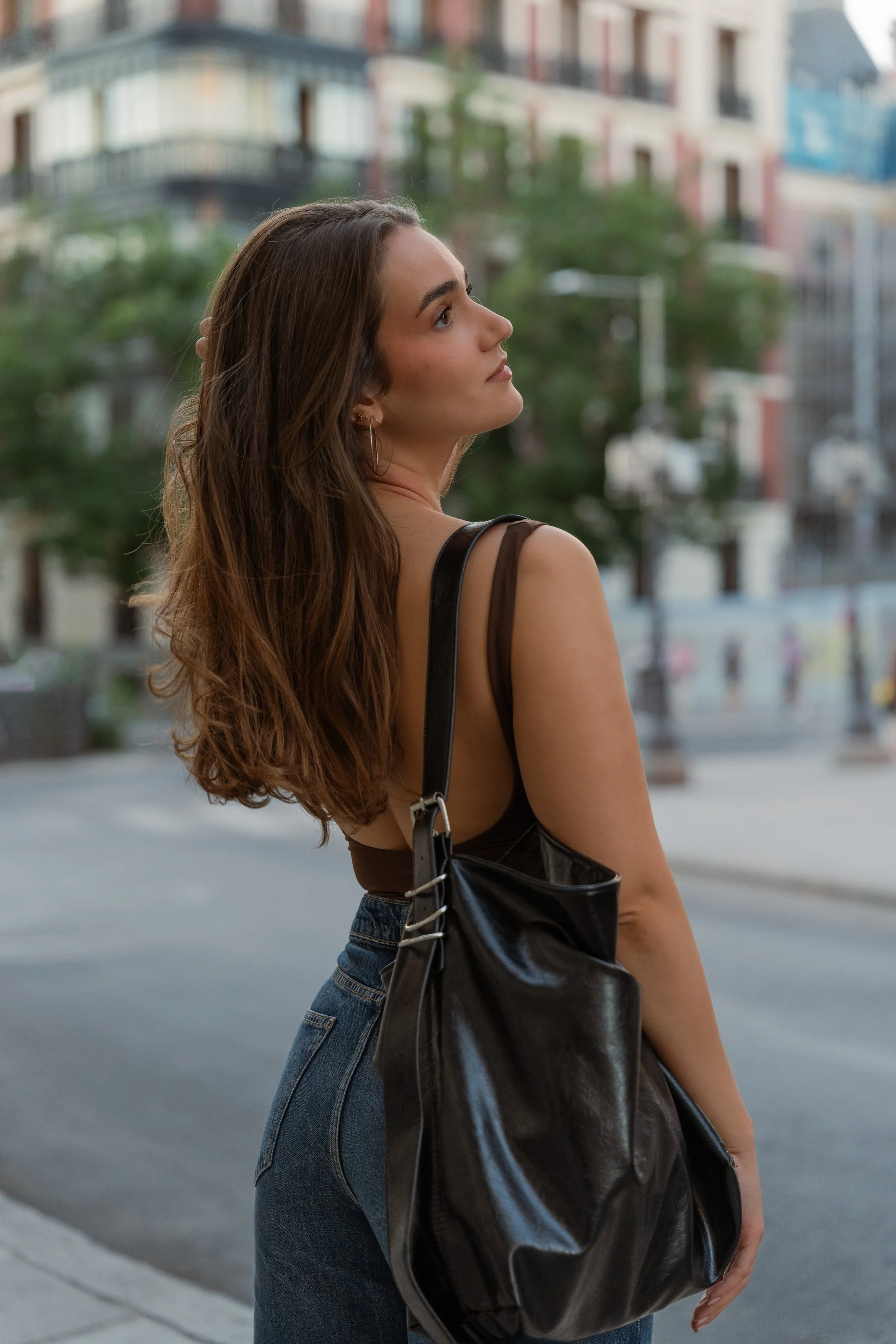 Young woman with long wavy brown hair wearing a black tank top and blue jeans, carrying a large black leather bag, standing on a city street, looking upward.