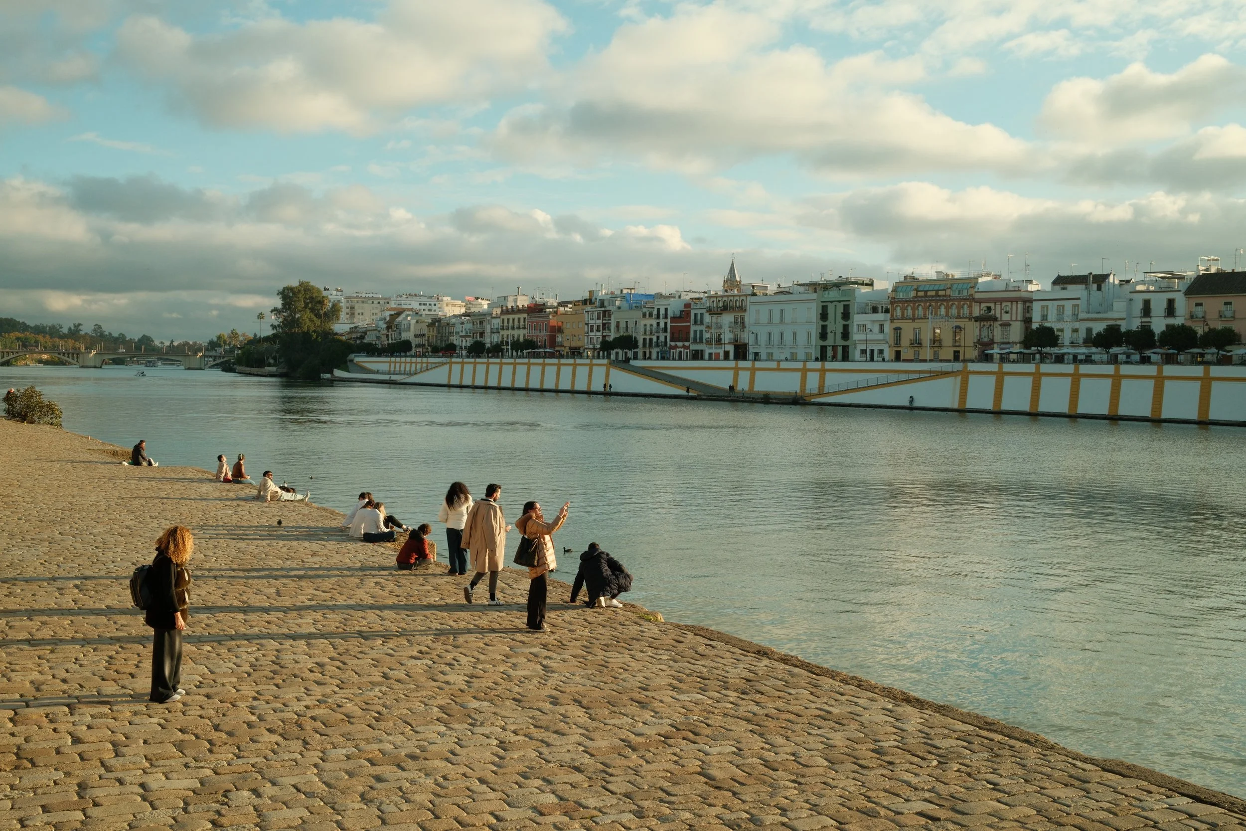 People sitting and walking along a cobblestone riverside promenade with a cityscape of colorful buildings across the water, under a partly cloudy sky.