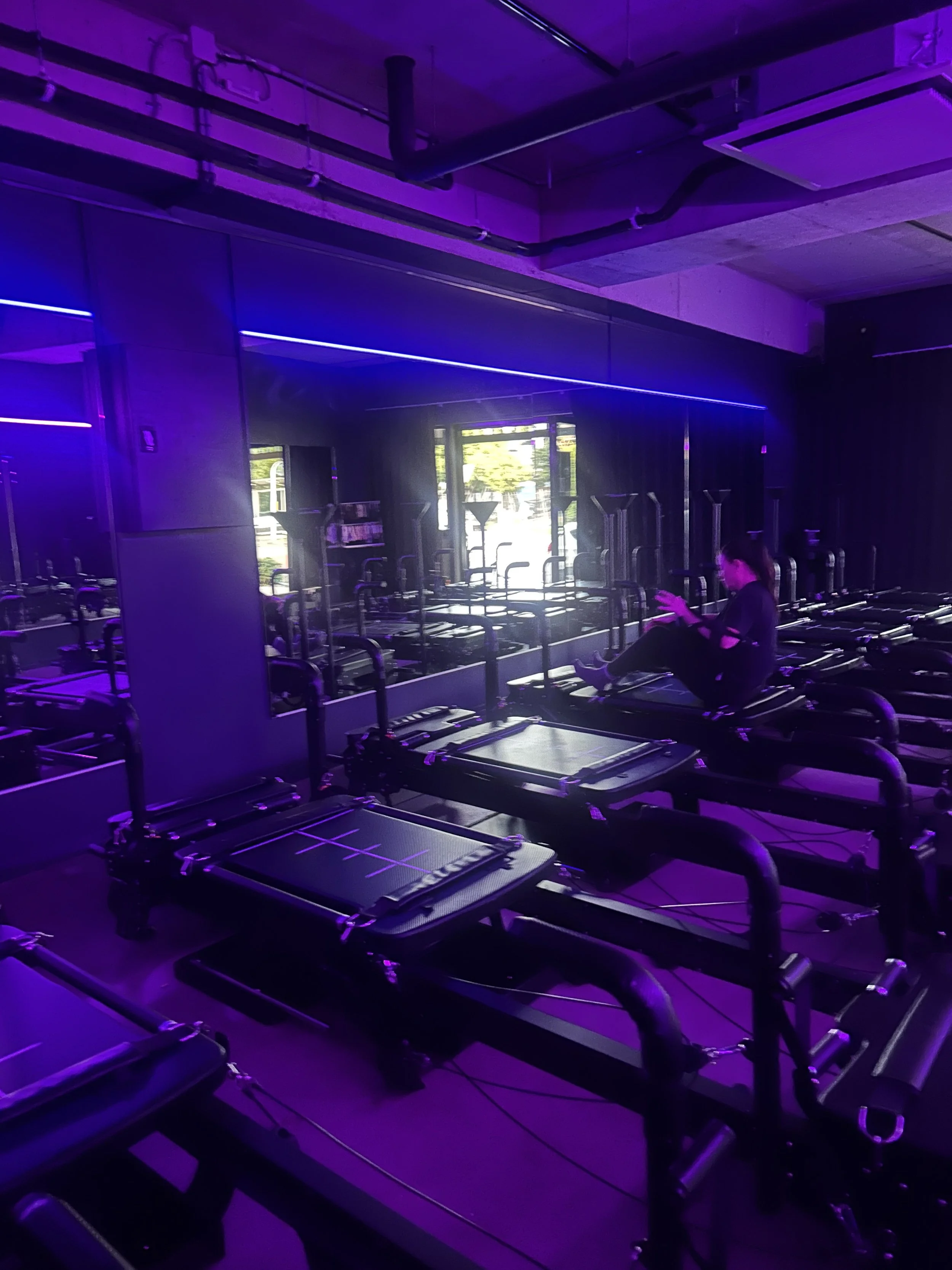 A dimly lit fitness studio with purple lighting, featuring multiple treadmills and a person sitting on one while looking at their phone. The room has large windows, mirrors, and exercise equipment.