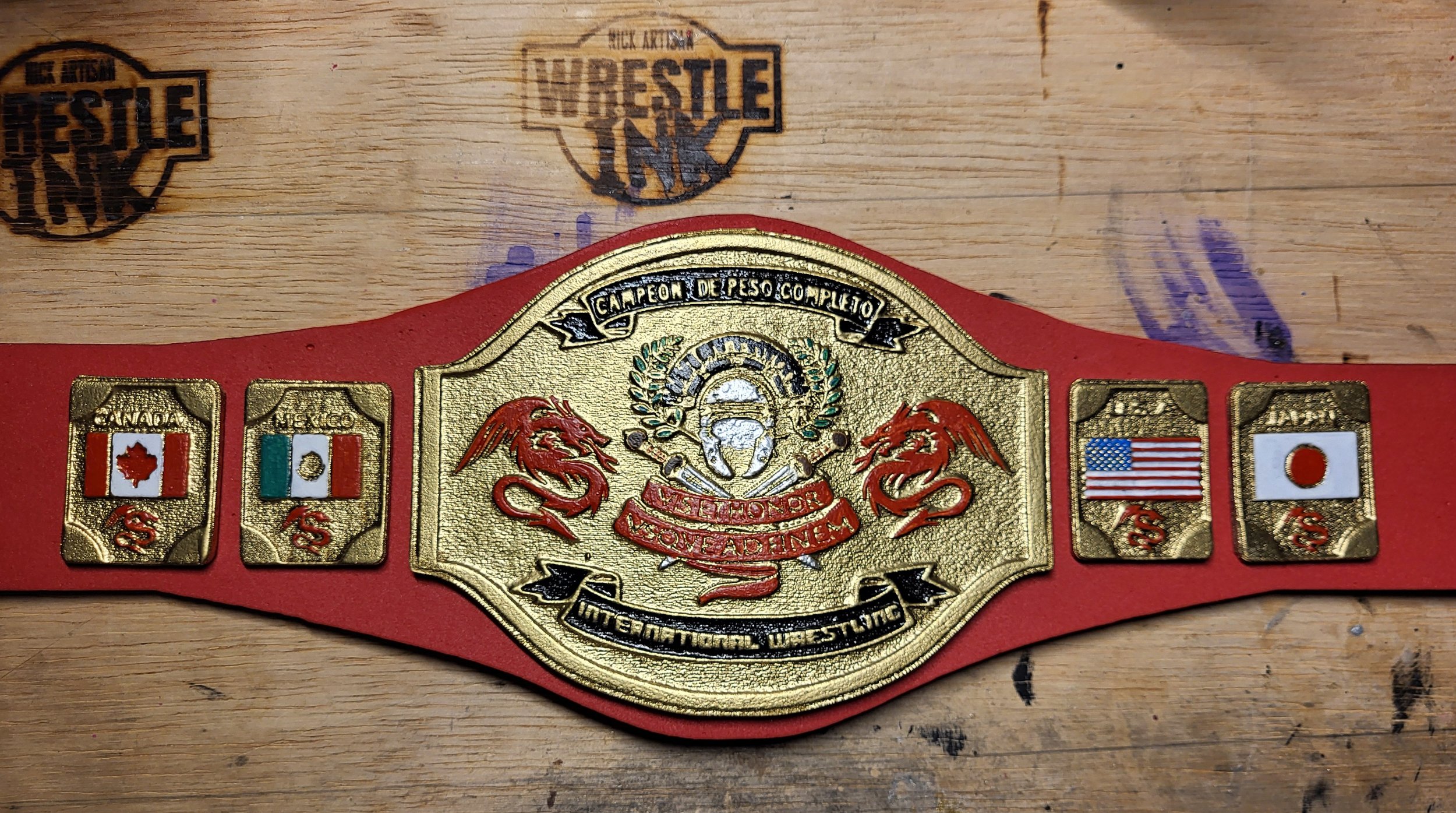 Red wrestling championship belt with gold plates, featuring flags of Canada, Italy, USA, and Japan, and text in Spanish about wrestling achievements, placed on a wooden surface.