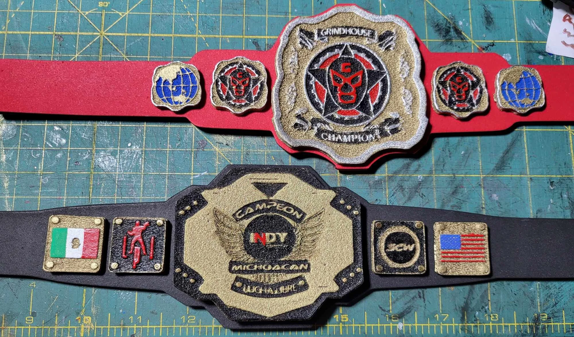 Two championship wrestling belts placed on a green cutting mat with yellow grid lines. The top belt has a red strap with various patches, including one with a black and red face and the words 'Groundothers Champion.' The bottom belt has a black strap