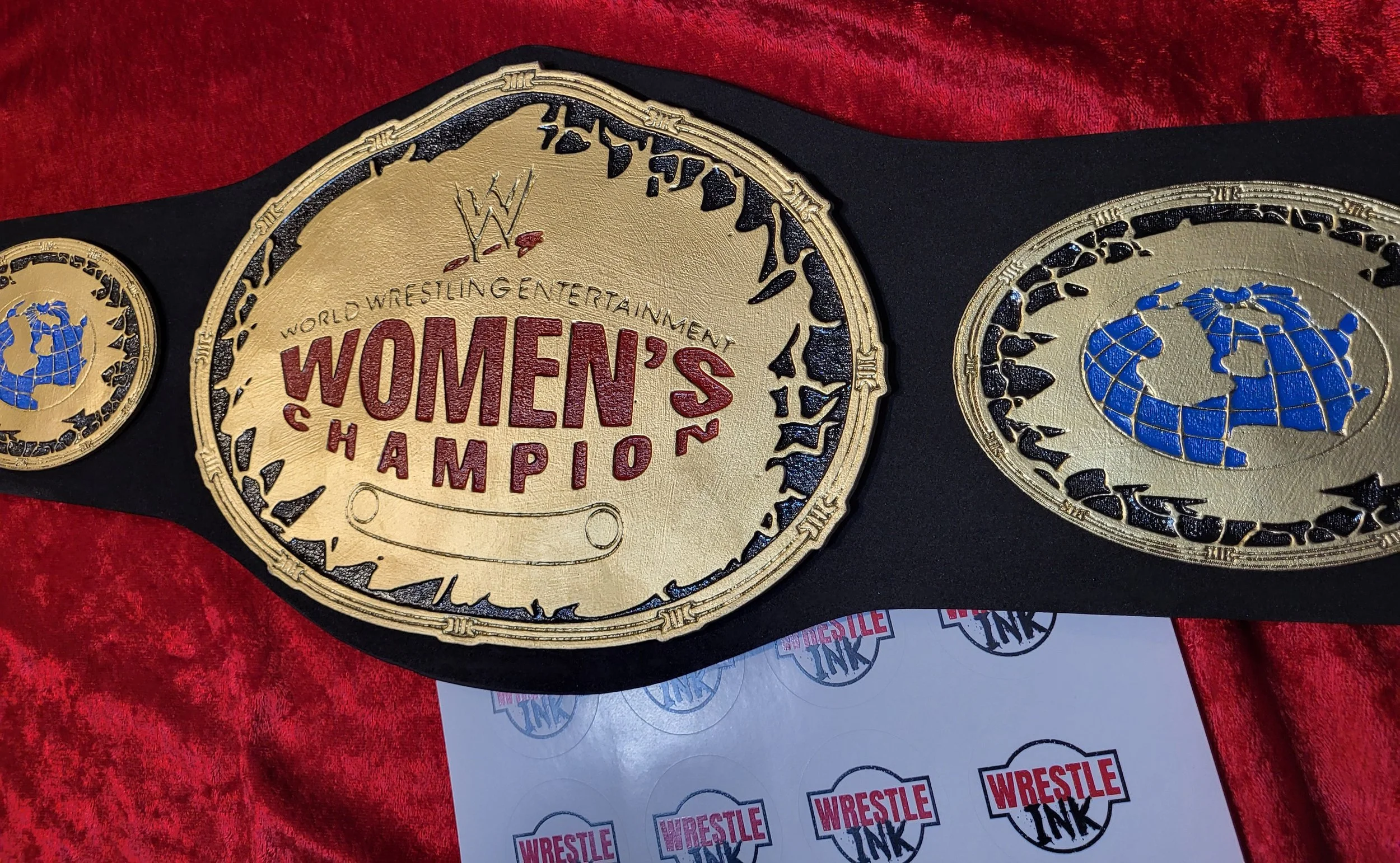 A wrestling championship belt with gold plates that read 'Women's Champion' and feature a blue globe emblem, placed on a red velvet surface with a sheet of stickers or labels nearby.