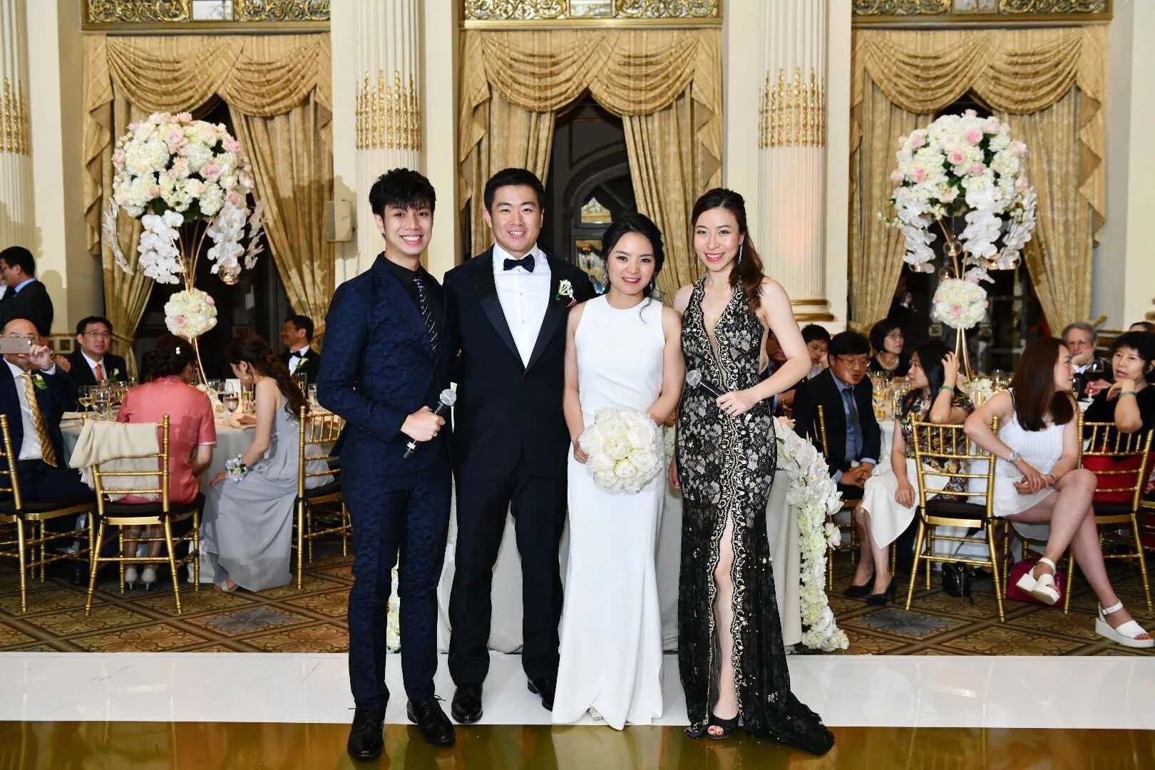 A group of five people dressed in formal attire at a wedding reception, standing in front of a decorated banquet hall with seated guests and floral centerpieces.