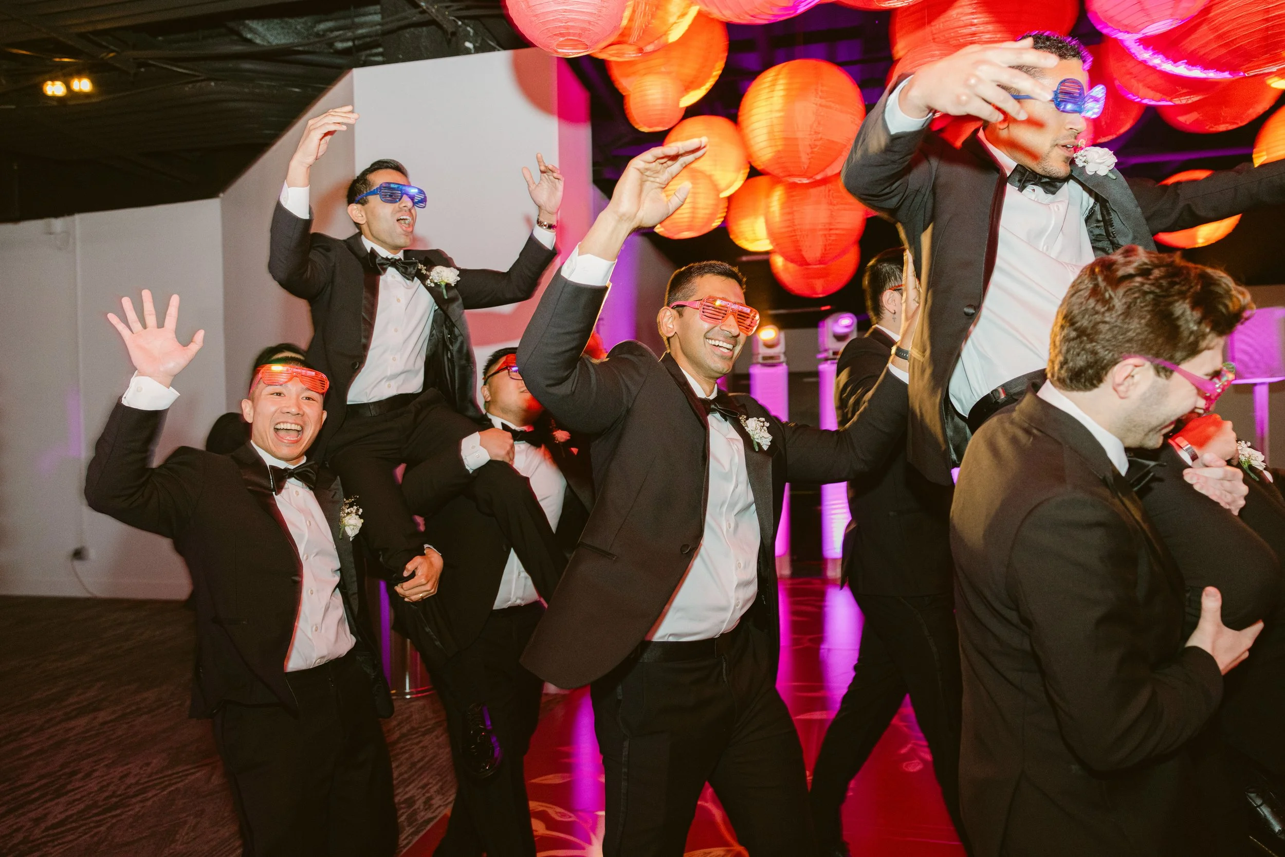 Men dressed in tuxedos wearing colorful glasses, performing a train dance at a party with red paper lanterns hanging from the ceiling.