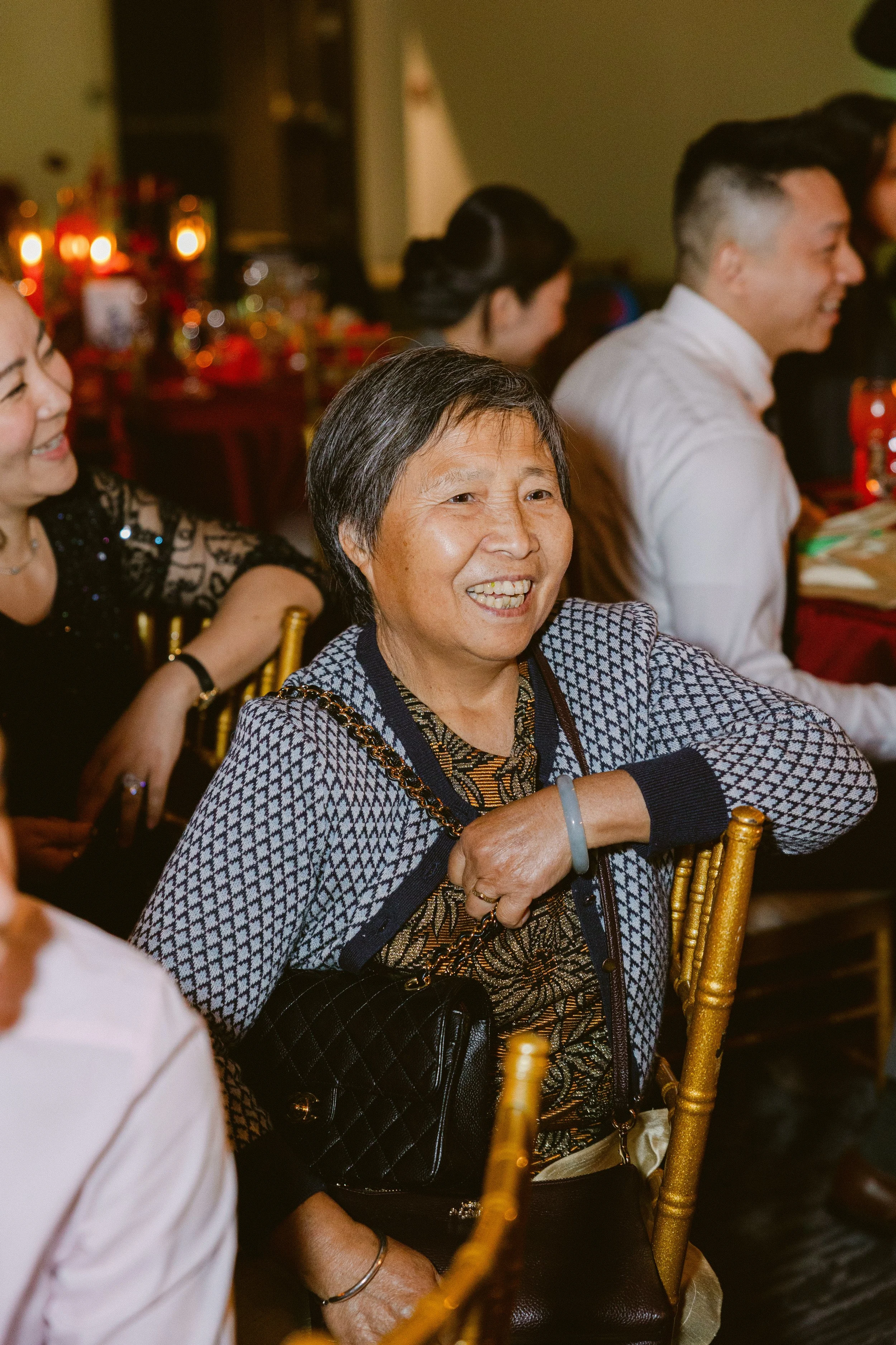 A woman smiling and sitting at a social gathering or celebration with other people, with tables and decorations in the background.