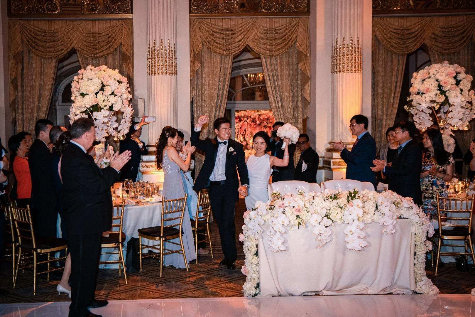 Couple at a wedding reception holding bouquets, surrounded by guests clapping and cheering, in a decorated venue with large floral arrangements and elegant drapes.