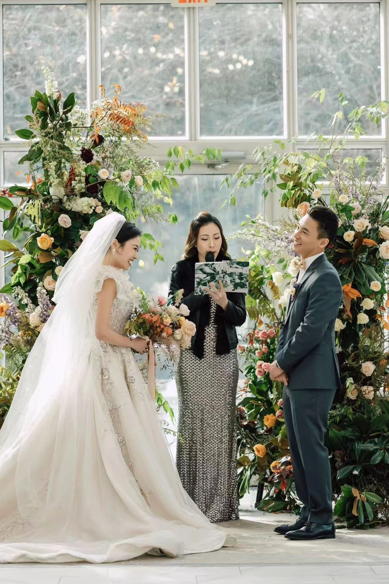 A bride and groom exchanging vows during a wedding ceremony, with an officiant reading from a book, surrounded by floral arrangements and greenery in a bright, indoor setting.