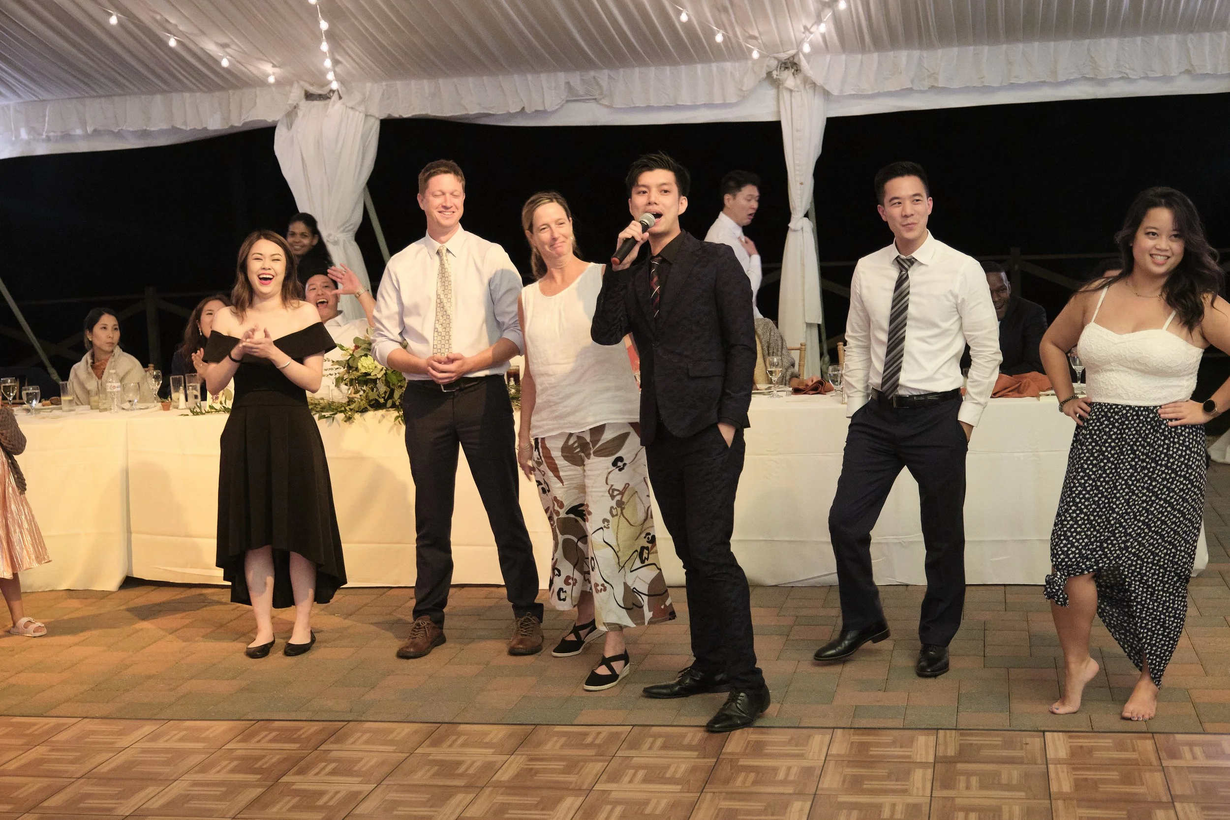 People standing and smiling at a celebration, with a man holding a microphone in the center, under a decorated tent, with a long table behind them.