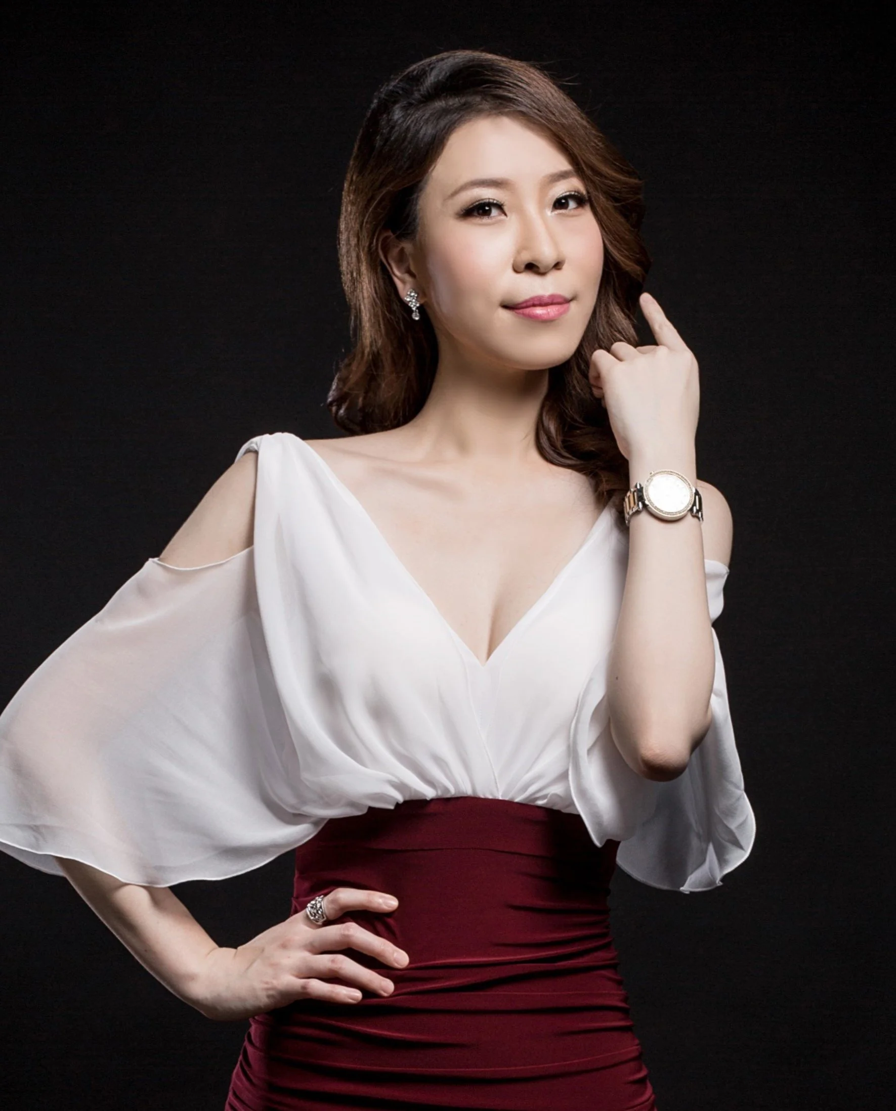 A woman with shoulder-length brown hair, wearing a white off-shoulder top and a burgundy skirt, posing with one hand on her hip and the other near her face against a black background.