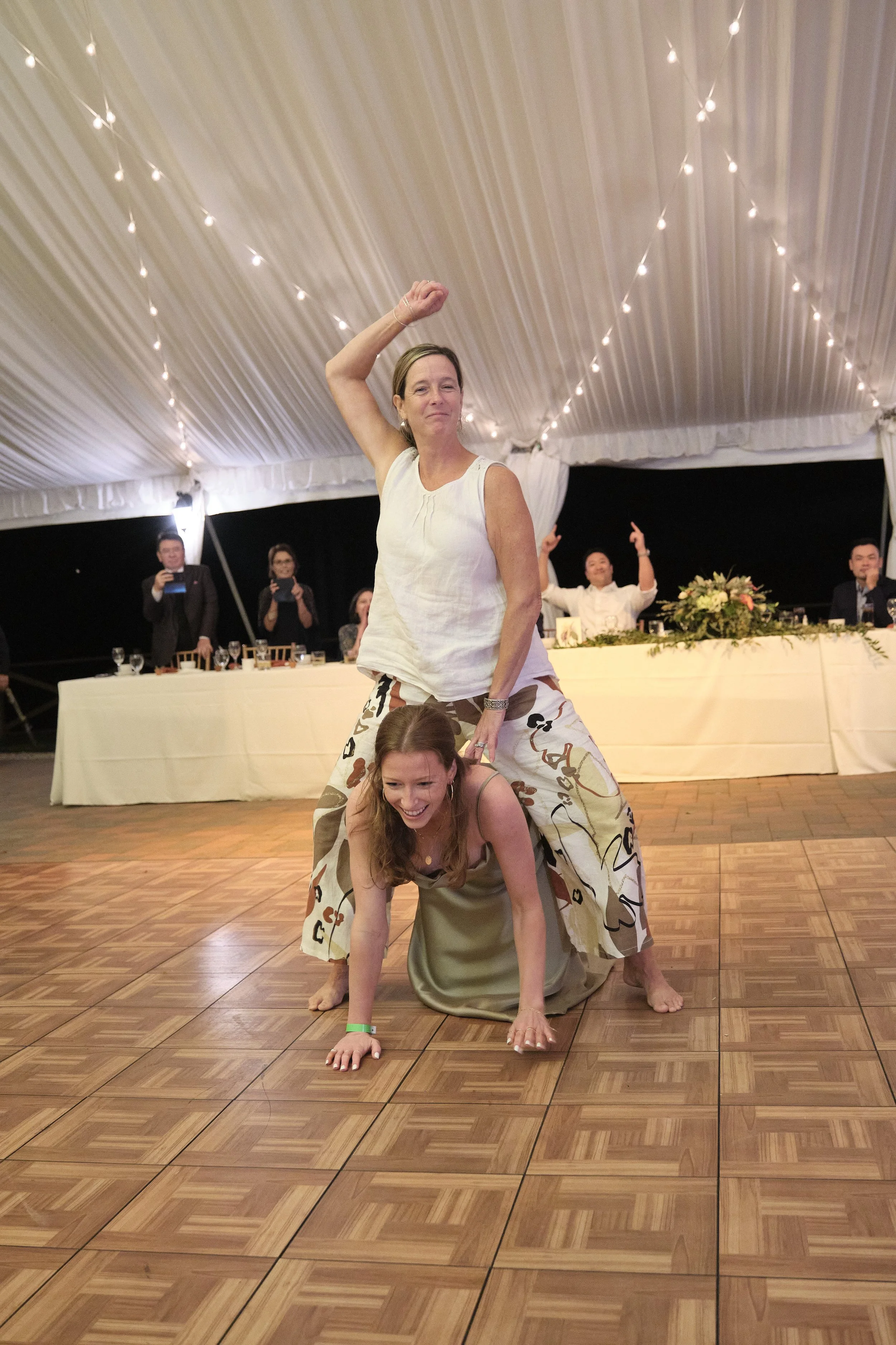 Two women performing a humorous dance routine at a formal event, with one woman on all fours and the other standing on her back with her arm raised, while guests watch and take photos in the background under a decorated tent ceiling.