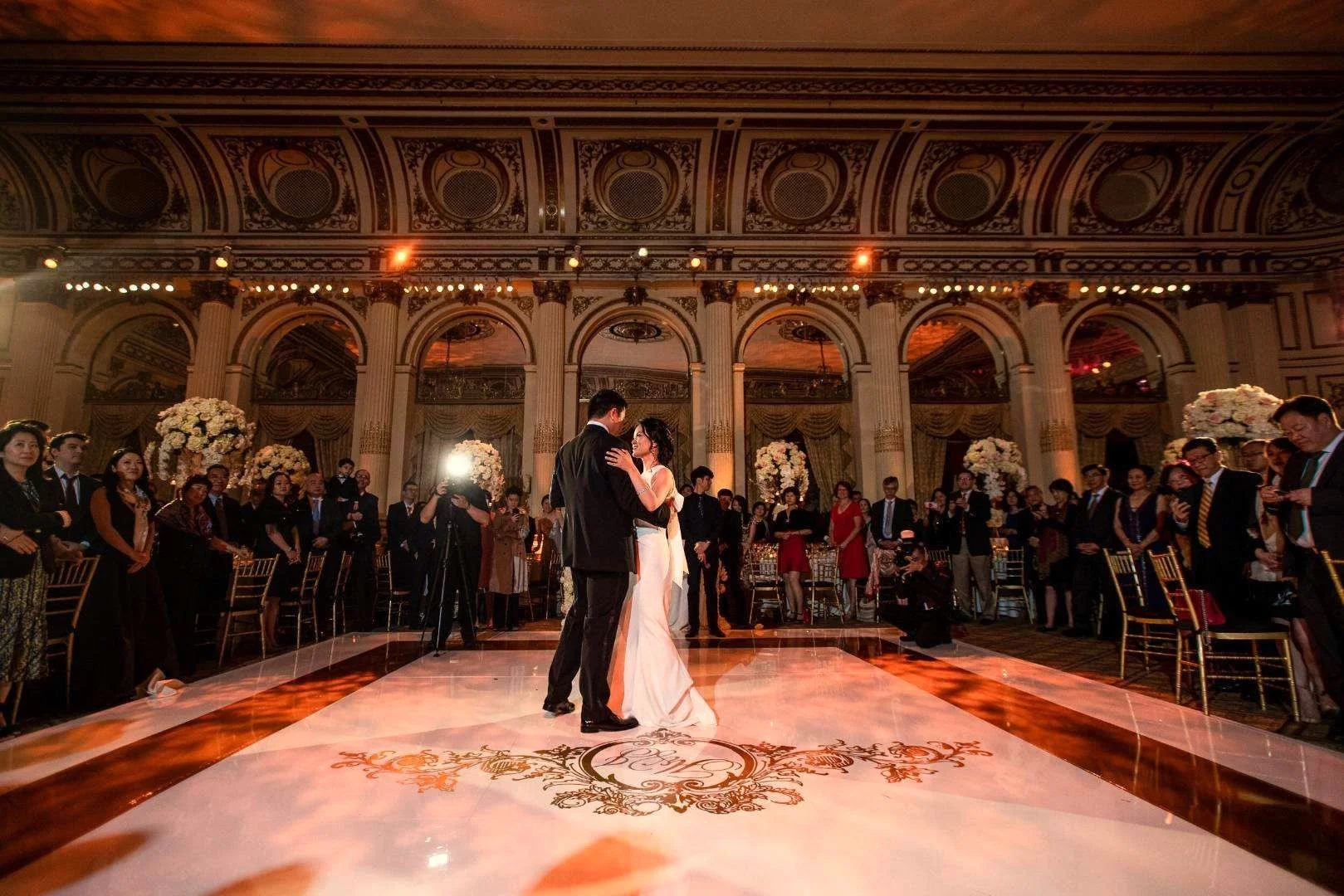 A bride and groom dancing at their wedding reception in a grand, ornate ballroom surrounded by seated and standing guests, with floral arrangements and decorative lighting.