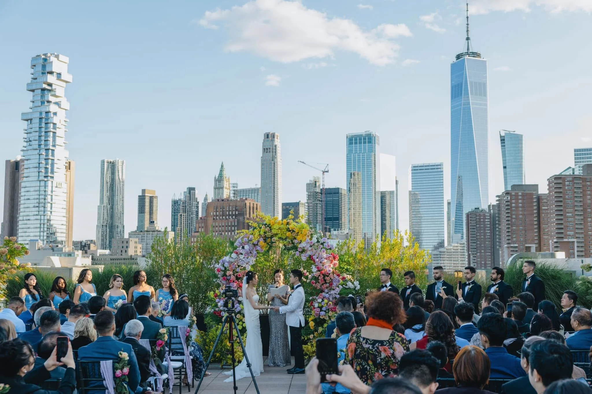 A wedding ceremony taking place outdoors with a city skyline in the background, including tall skyscrapers and the One World Trade Center, with guests seated, a floral arch, and the couple exchanging vows.