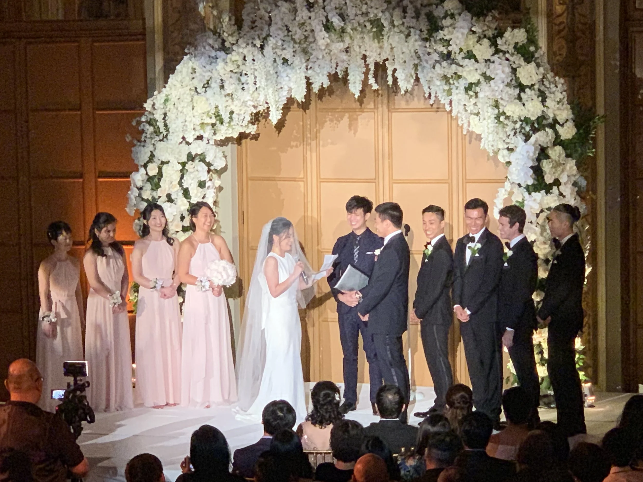 A wedding ceremony with the bride in a white dress and veil, and the groom in a suit, exchanging vows under a floral arch. Bridesmaids in pink dresses and groomsmen in black suits are standing nearby, with guests seated in front watching the event.