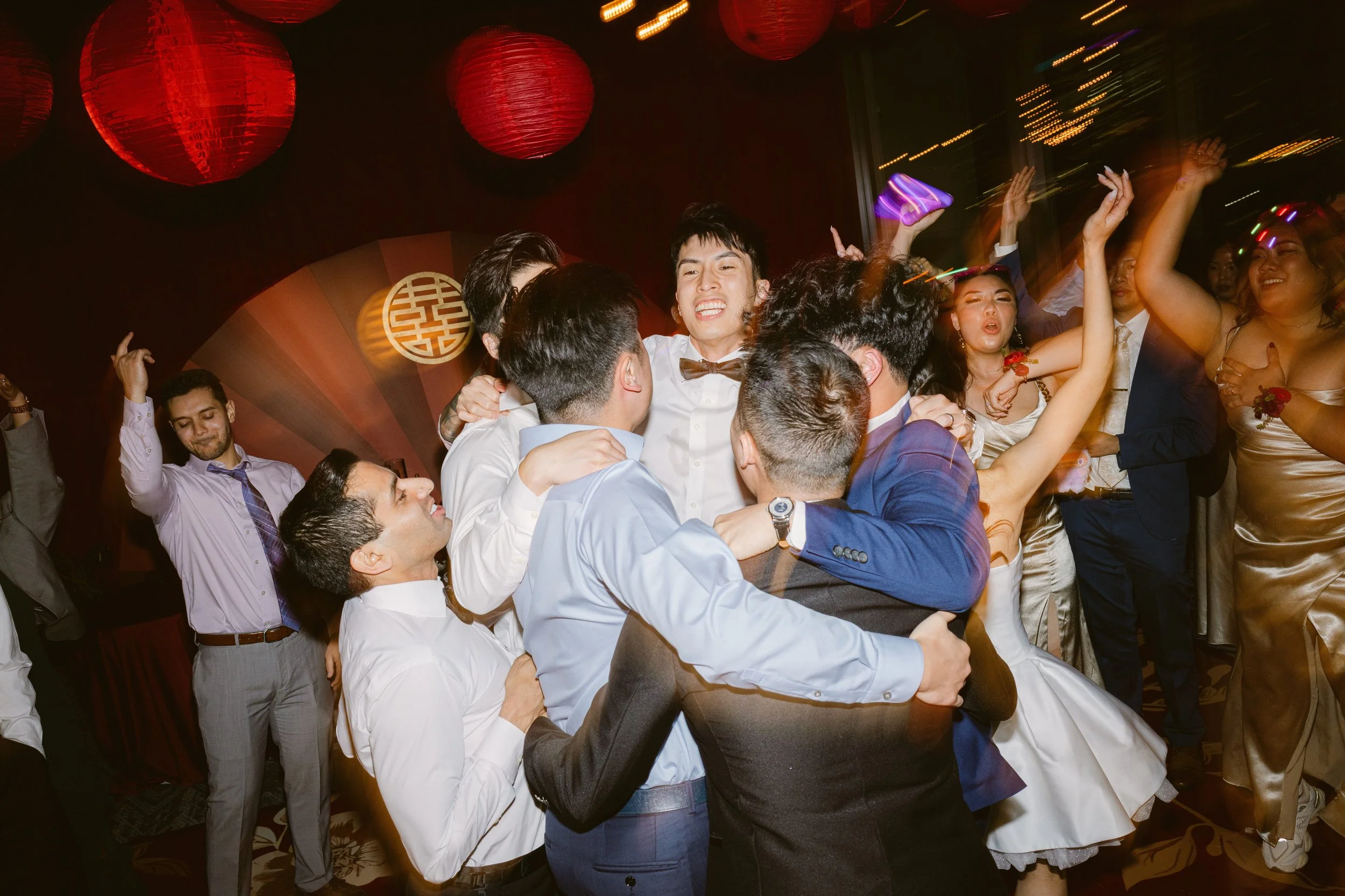 Group of people dancing and celebrating at a party or wedding reception, with some people hugging and smiling, in a decorated venue with red lanterns and Asian-themed décor.