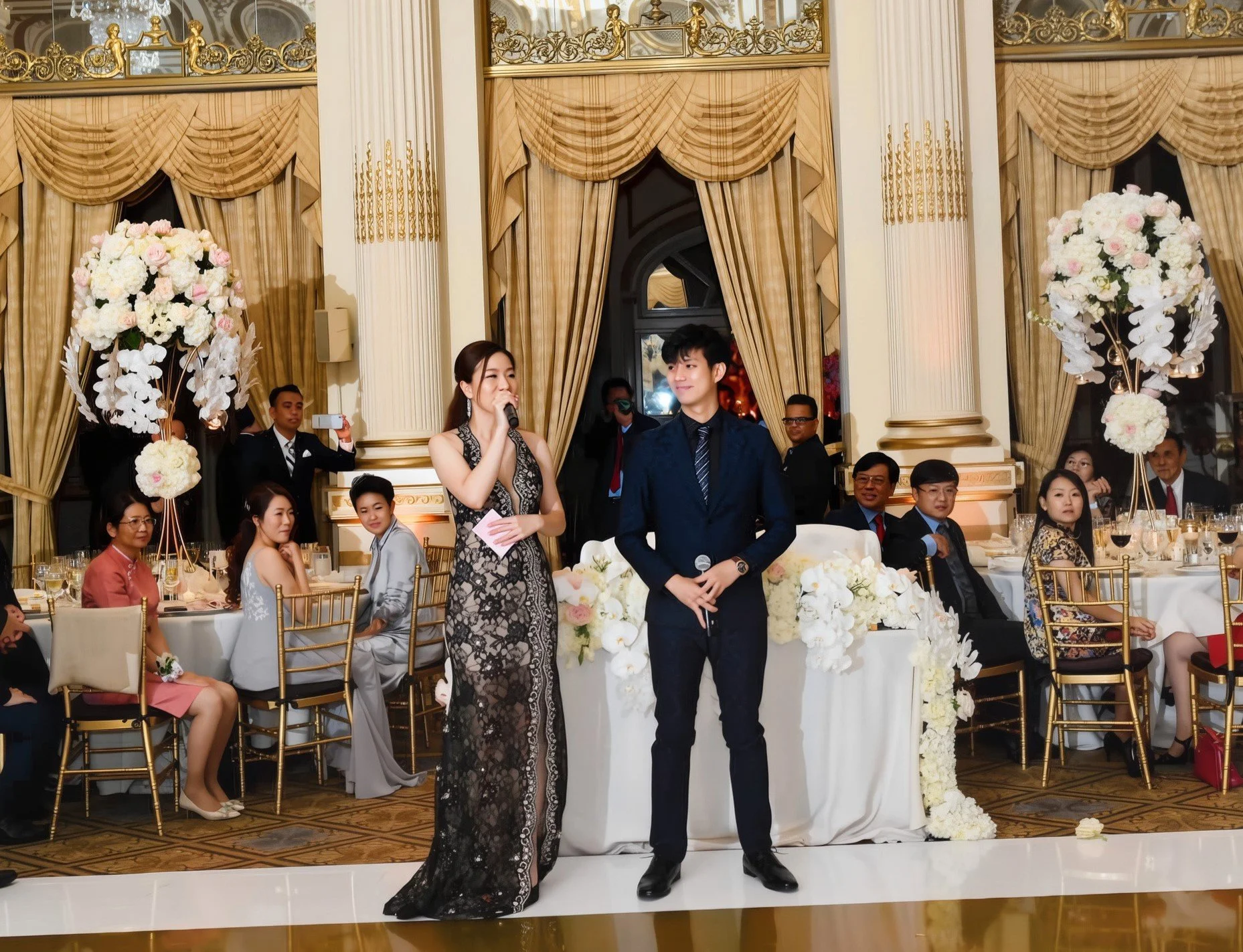 A wedding reception with a woman and a man standing in front of a decorated table, surrounded by seated guests in elegant attire, ornate gold and cream decor, large floral arrangements, and gold chairs.