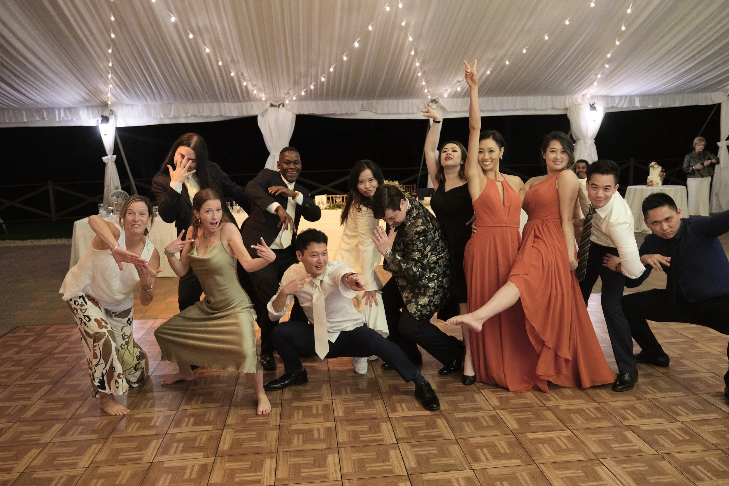 Group of people at a celebration, dancing and posing on a dance floor under string lights in a decorated tent.