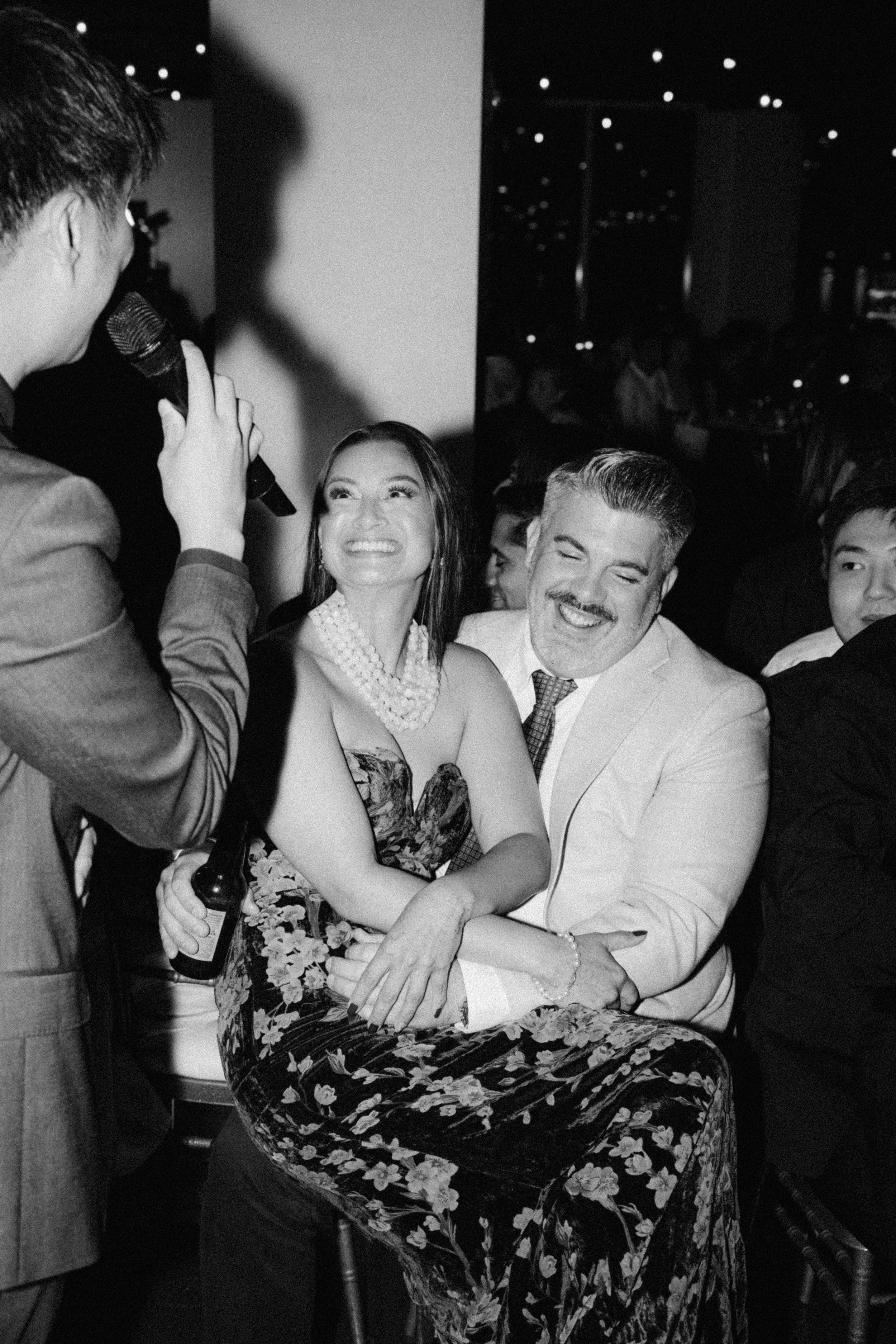 A man and woman laughing and hugging at a social event, with a person speaking into a microphone in the foreground.