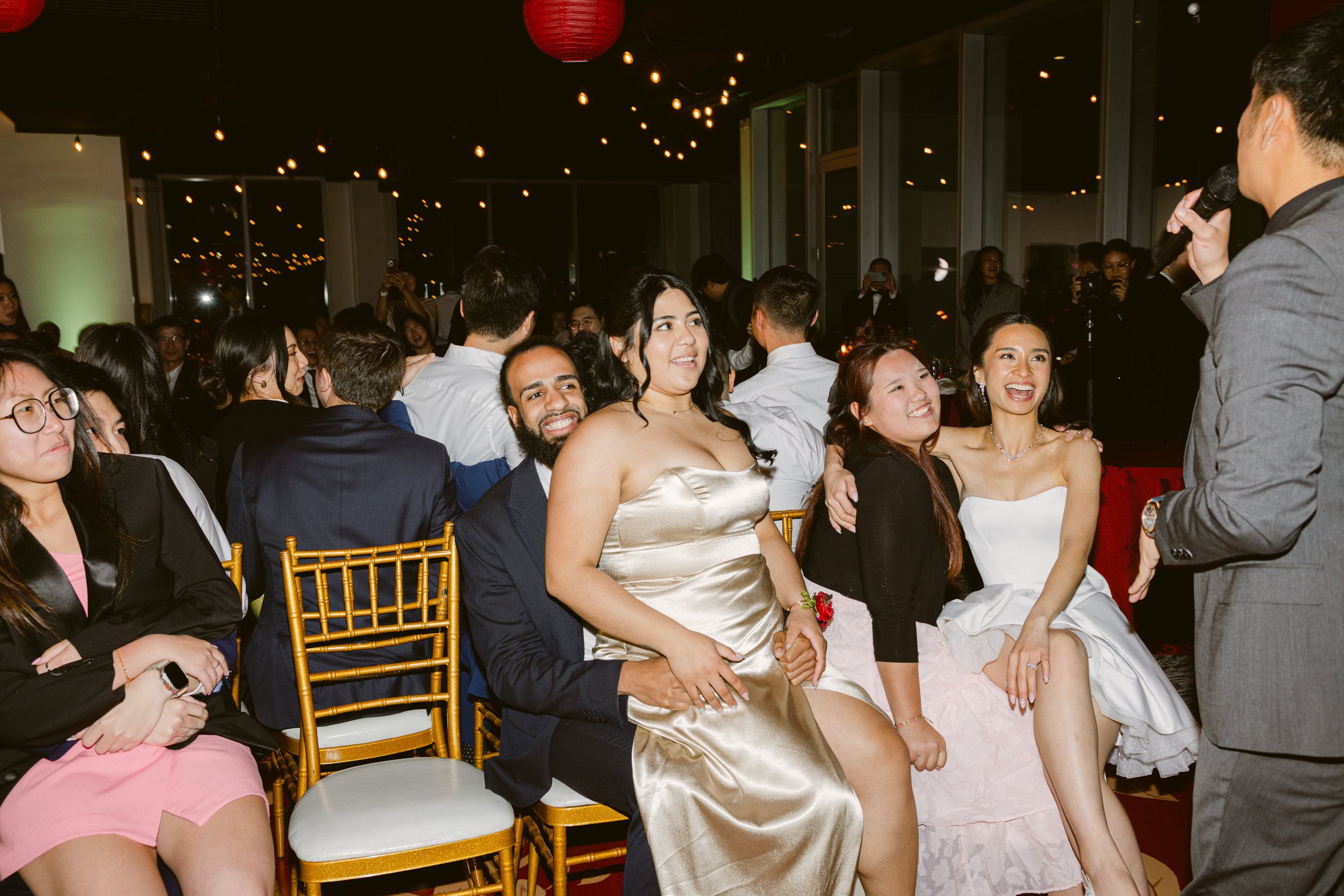 People seated at a formal event, with a man speaking into a microphone. Some guests are smiling and enjoying the moment.