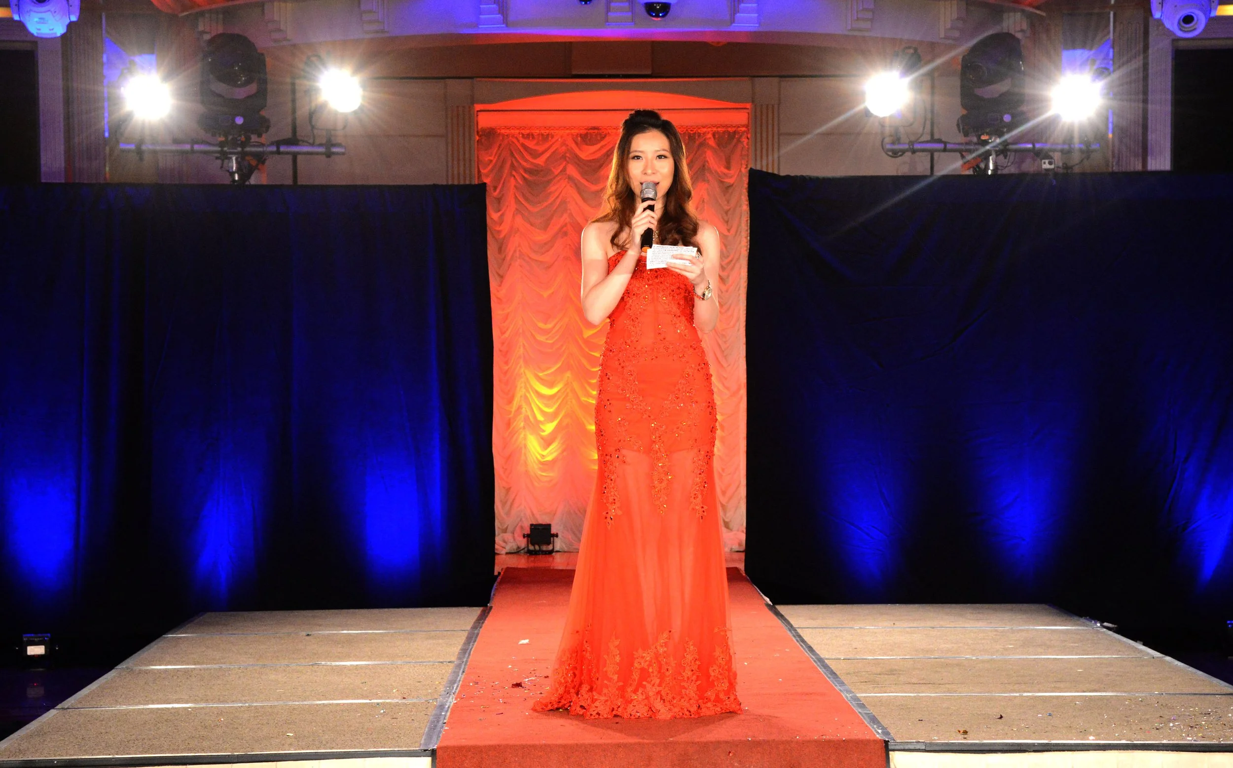 A woman in a red evening gown standing on a stage, holding a microphone and notes, with stage lights shining behind her.