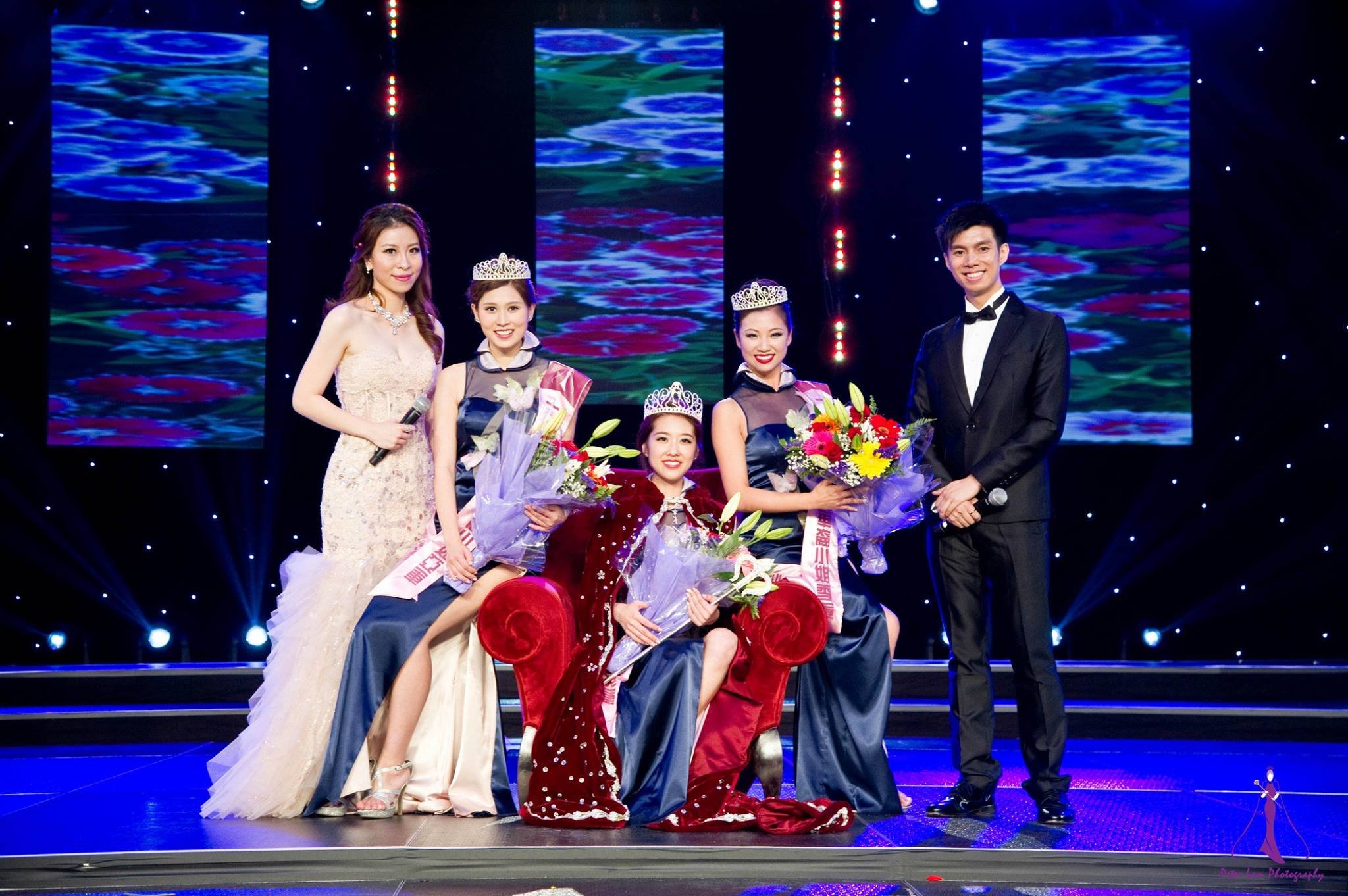 A beauty pageant awards ceremony on stage with five people: three women wearing crowns and sashes holding bouquets, a woman in a long white gown, and a man in a tuxedo holding a microphone. The woman sitting in the center is the winner, and the group