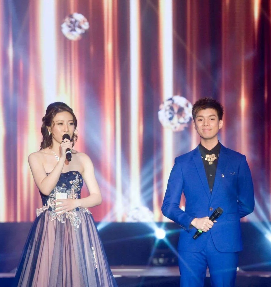 Two young people, a woman in a strapless evening gown and a man in a blue suit, stand on a stage with a vibrant, curtain-like backdrop. The woman is holding a microphone and appears to be speaking, while the man is smiling and holding a microphone.