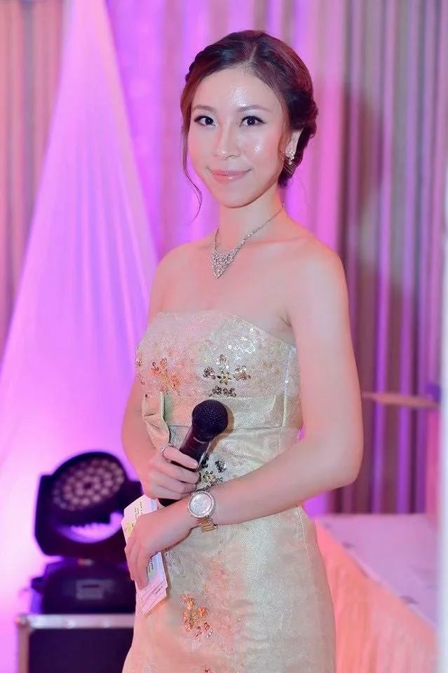 A woman in an elegant, strapless, gold, floral-patterned dress holding a microphone, standing in a decorated venue with pink and purple lighting.