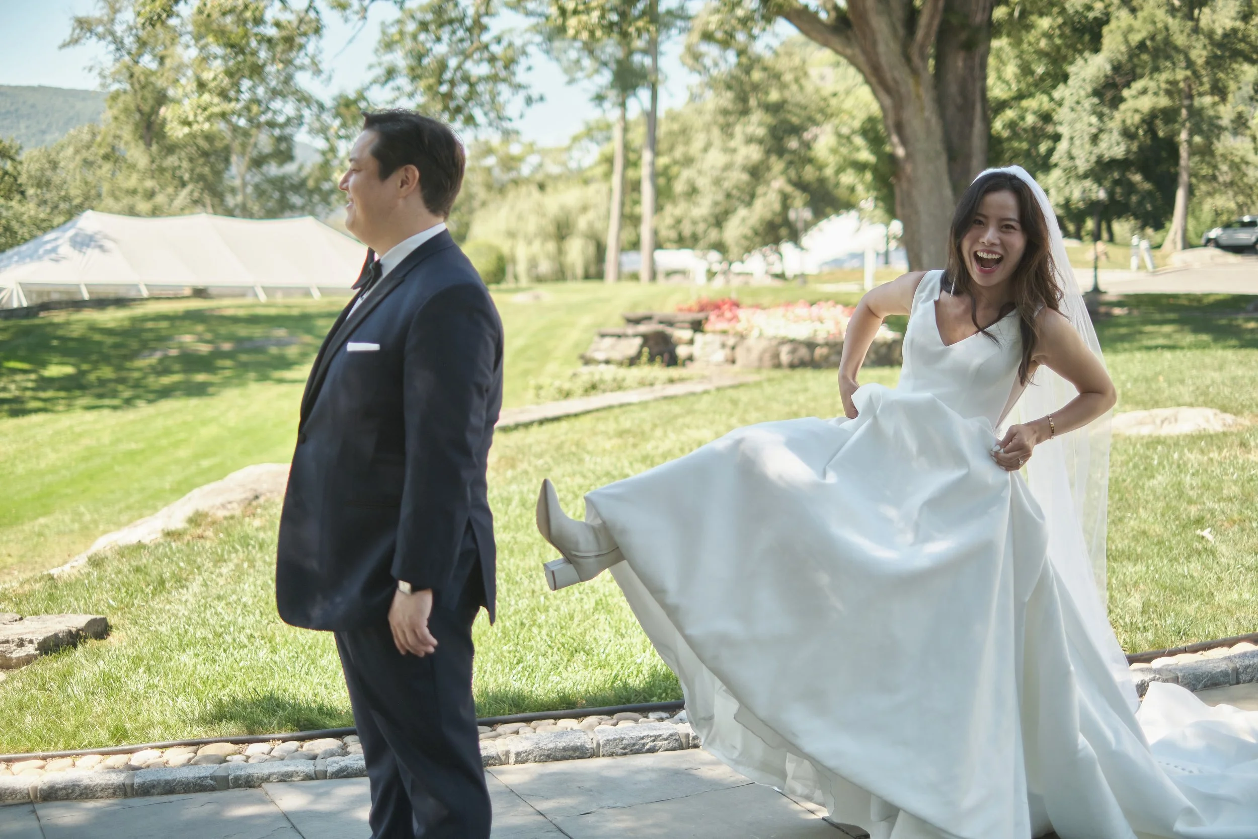 A bride in a white wedding dress and veil is holding up her dress and laughing, standing behind a groom in a navy suit outdoors in a park with trees and grass.