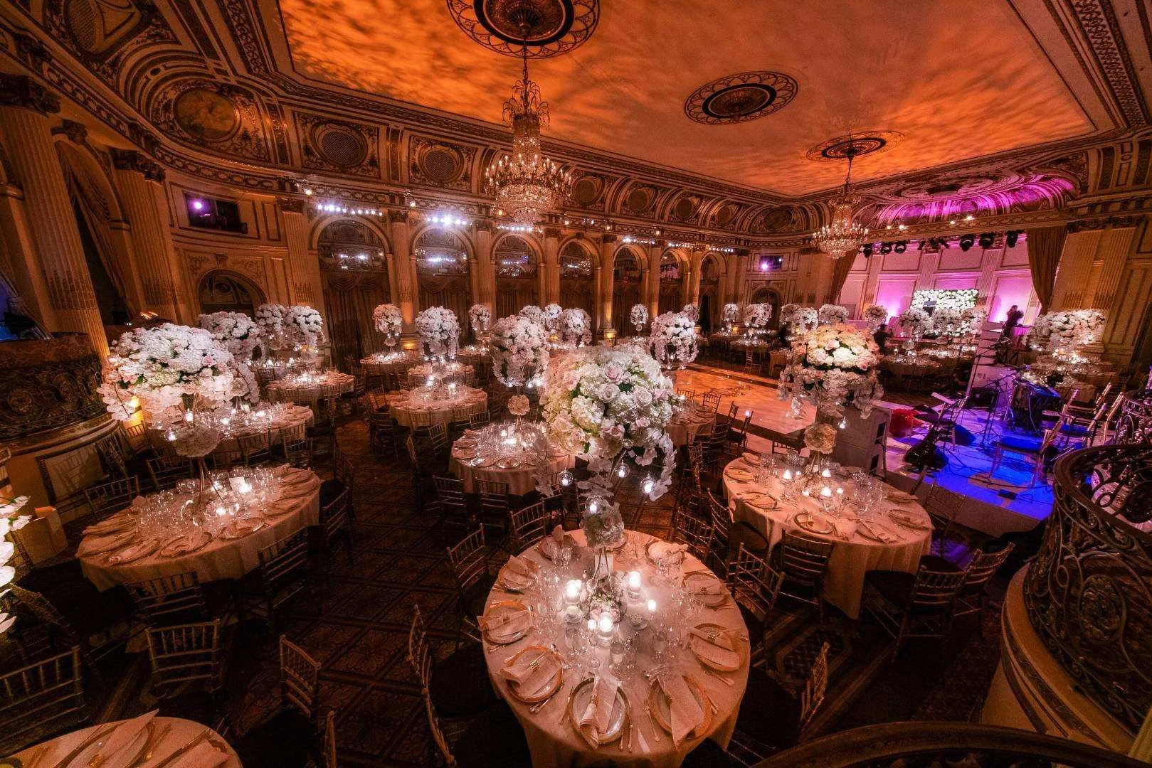 Elegant event hall decorated with numerous tall floral centerpieces featuring white flowers on round tables with pink tablecloths, set for a formal occasion with glassware and plates, with a stage and musical instruments at the front and ornate ceilings with chandeliers and pink lighting.