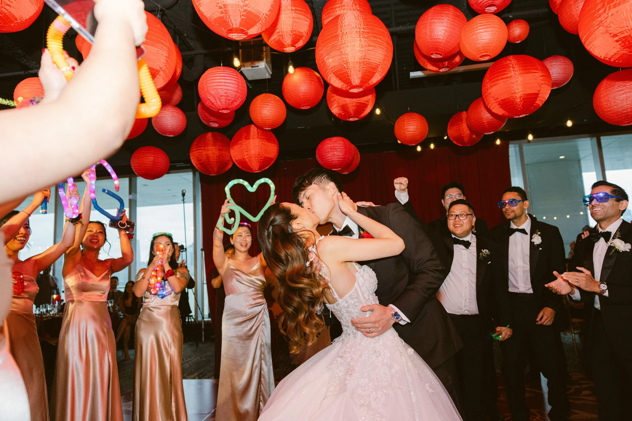 A bride and groom kiss during their wedding reception, surrounded by friends holding glowing signs and cheering, with red lanterns hanging overhead.